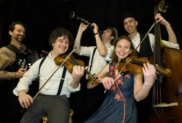 Five musicians, four men and one woman, playing string and wind instruments, performing on stage with joyful expressions.