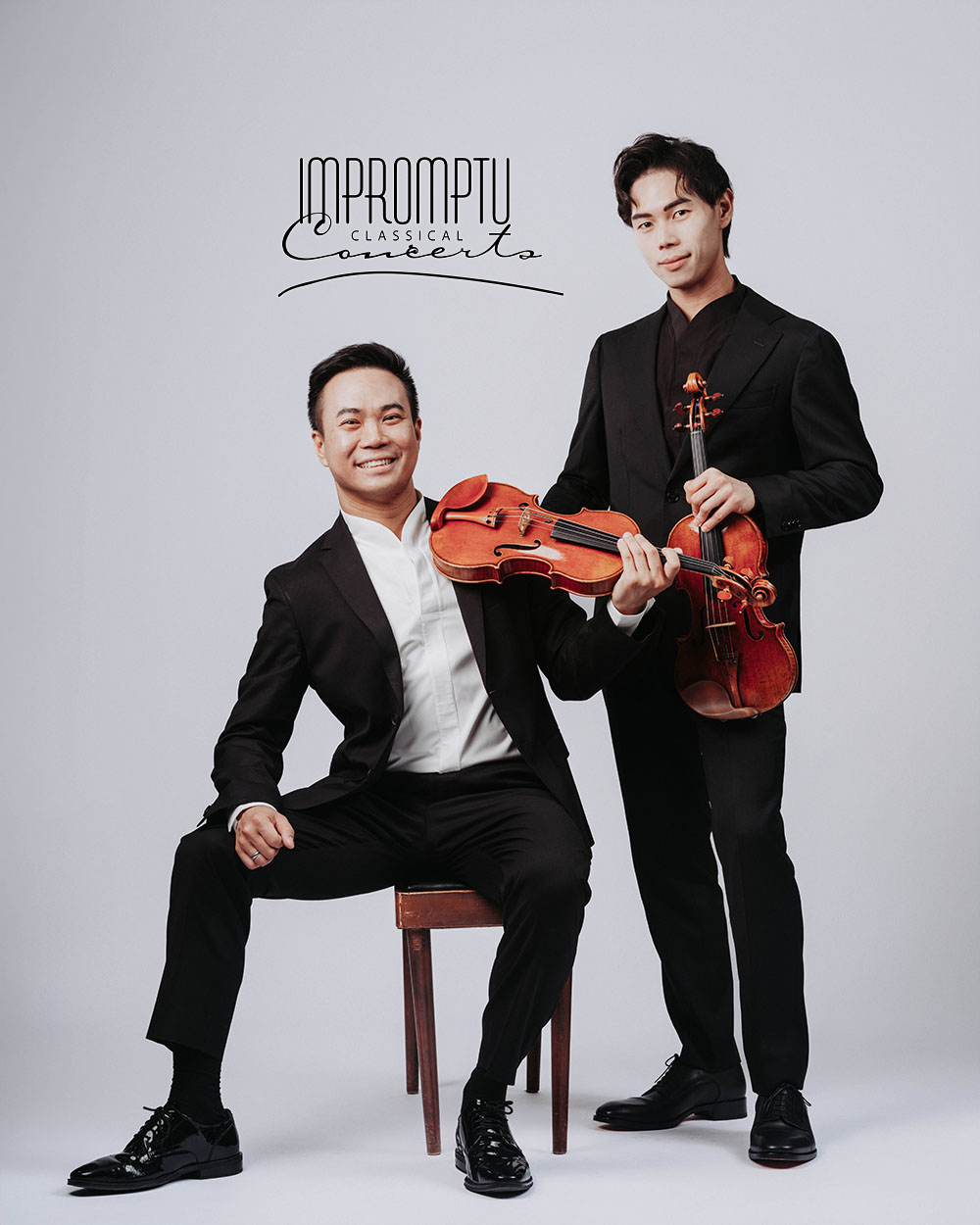 Two men in black suits holding violins, one seated and smiling, the other standing and holding his violin, against a plain gray background with the text 'Impromptu Classical Concerts'.