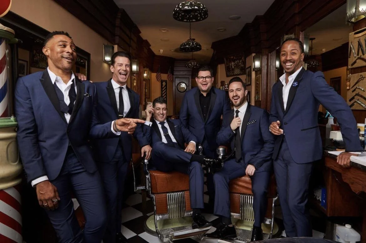 Group of six men in suits laughing and smiling indoors at a barbershop or barbershop-themed setting.