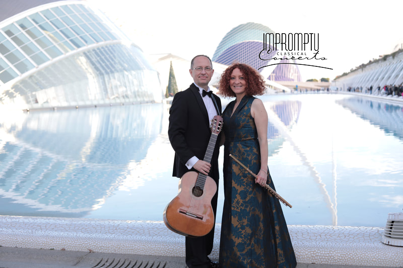 A man in a tuxedo holding a guitar and a woman in a gown holding a baton, standing in front of an architecturally unique building with water reflecting beside them.