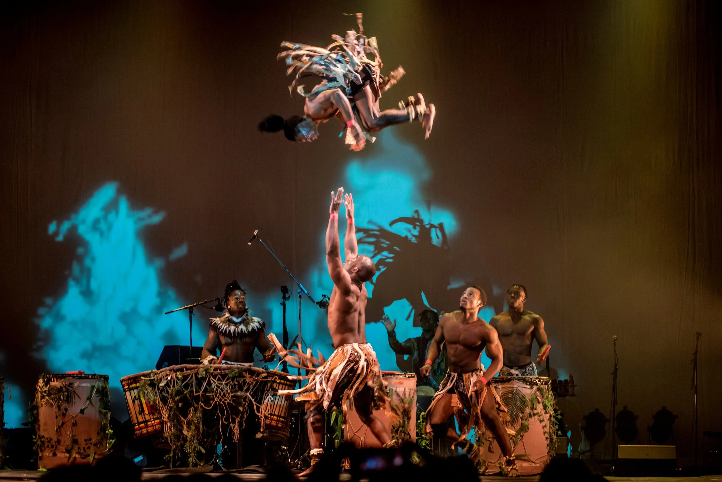 Performance on stage with natives wearing tribal costumes, drumming, and dancing, with a person floating in mid-air.