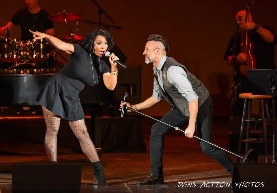 A woman singing into a microphone while a man crouches with another microphone on a stage with musical instruments in the background.