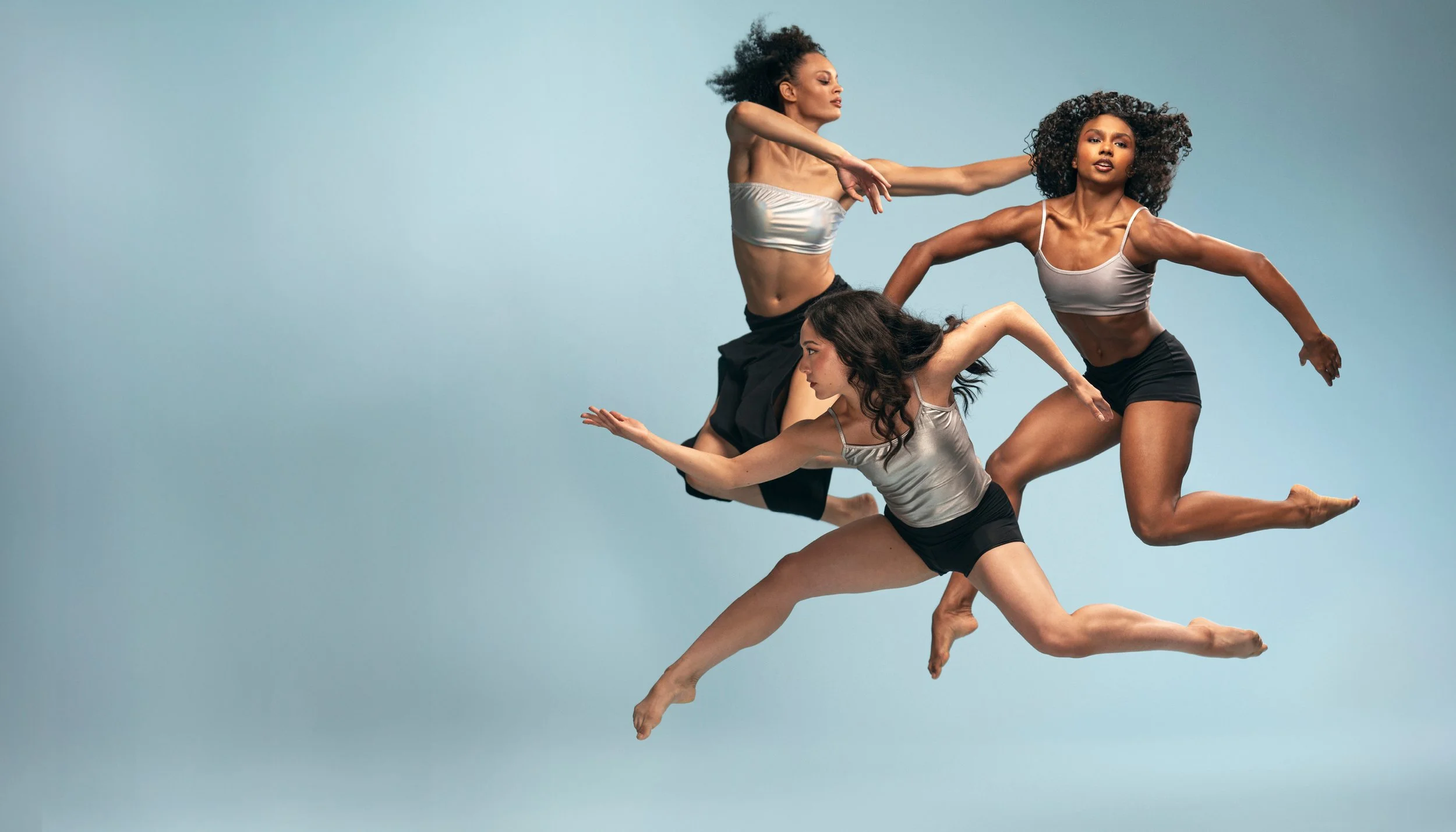 Three women in athletic attire performing a coordinated dance or pose against a light blue background.