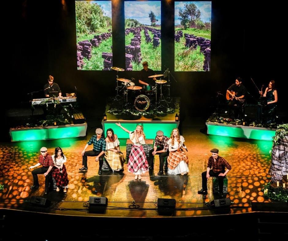 A group of performers on stage during a theater production, with a backdrop of three screens displaying outdoor scenery with trees, grass, and a stone path. The stage features musical instruments, including a keyboard, drums, and guitars, with some actors dressed in vintage clothing, suggesting a period setting.