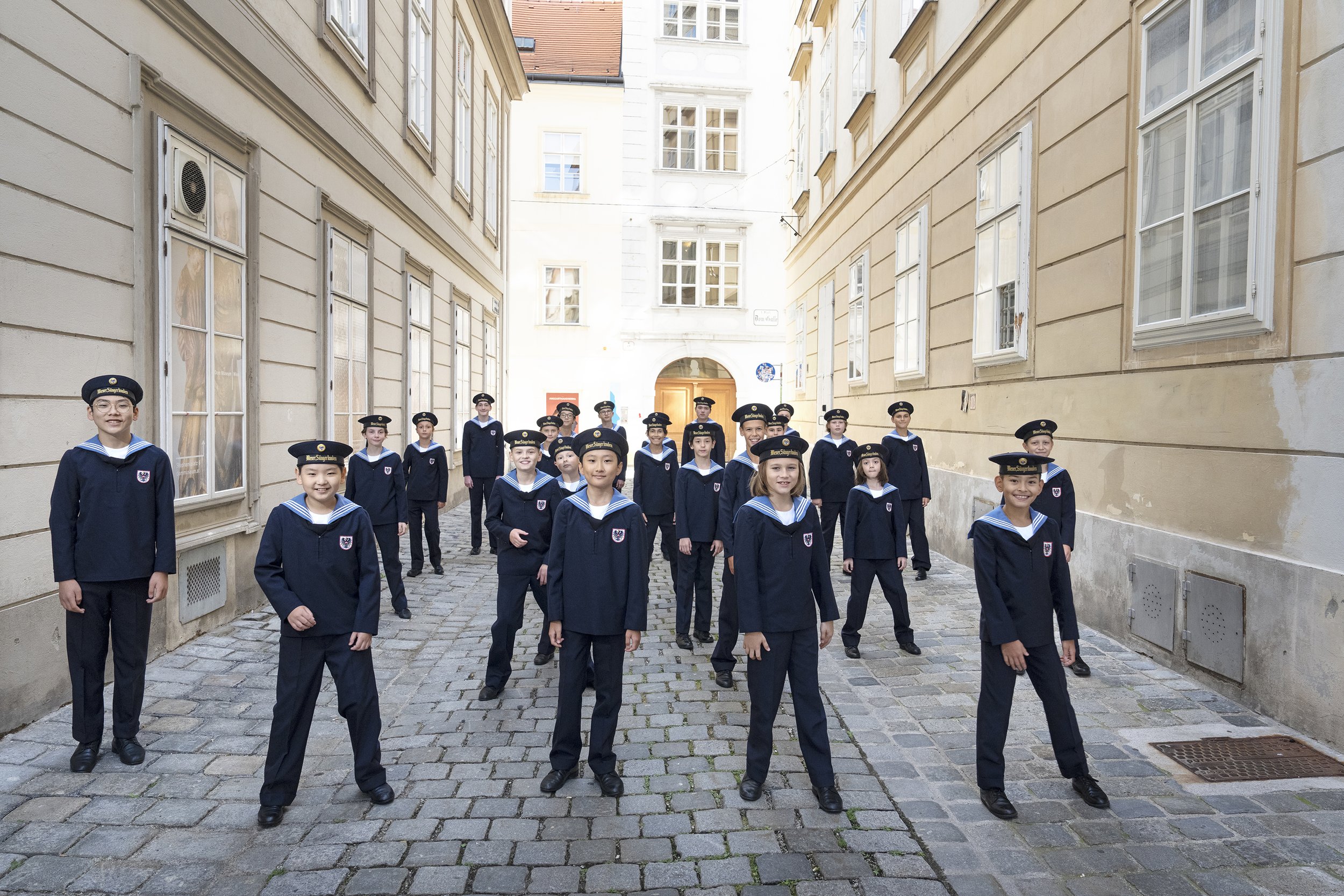 Four children in sailor outfits jumping in the air in front of a white building with green bushes and grass
