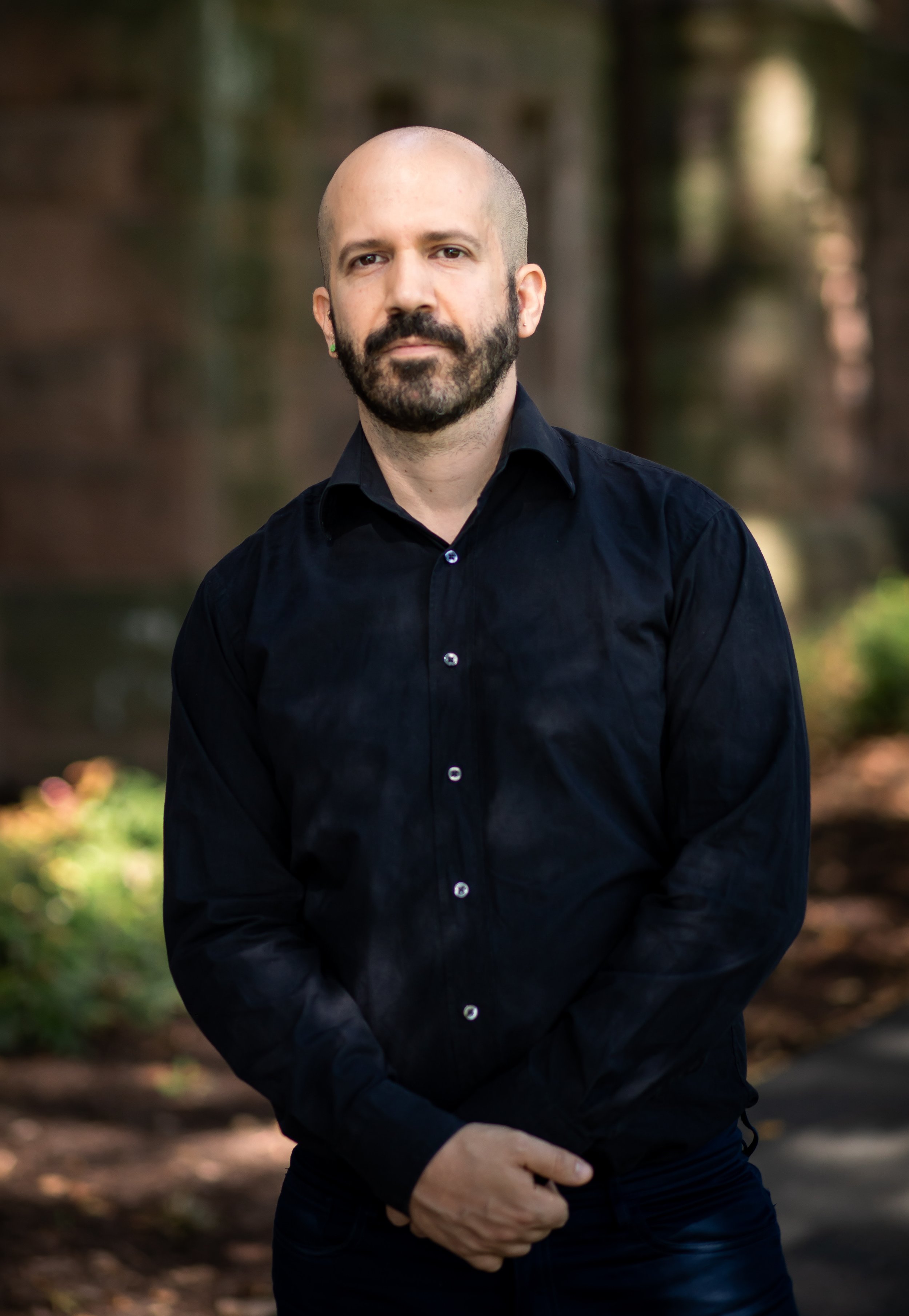 A bald man with a beard wearing a black button-up shirt stands outdoors with blurred nature trees and sunlight in the background.