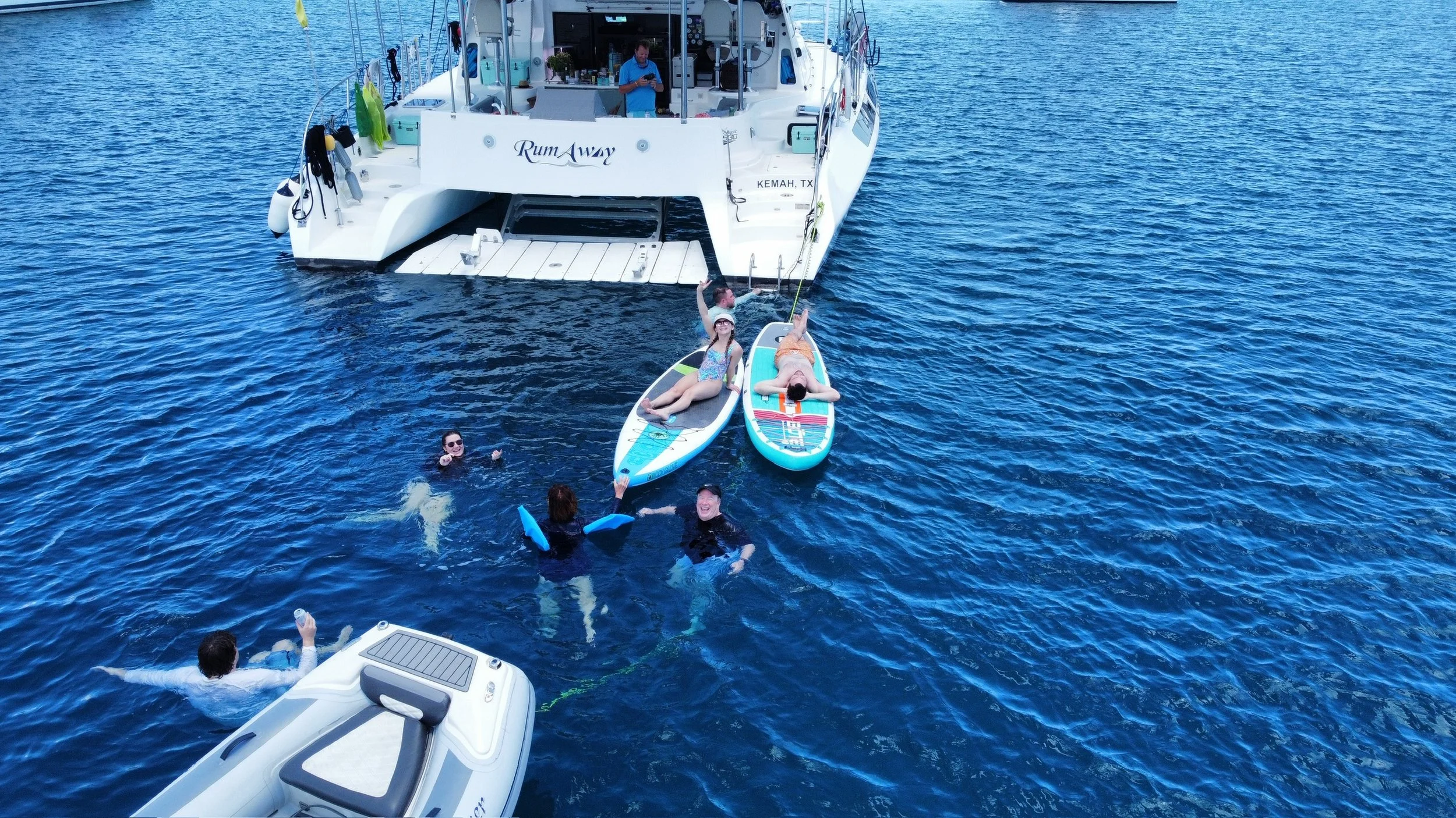 People swimming and relaxing in the water near a large white yacht named 'Rum Away' docked on a body of water, with some standing on paddleboards and others floating with flotation devices.
