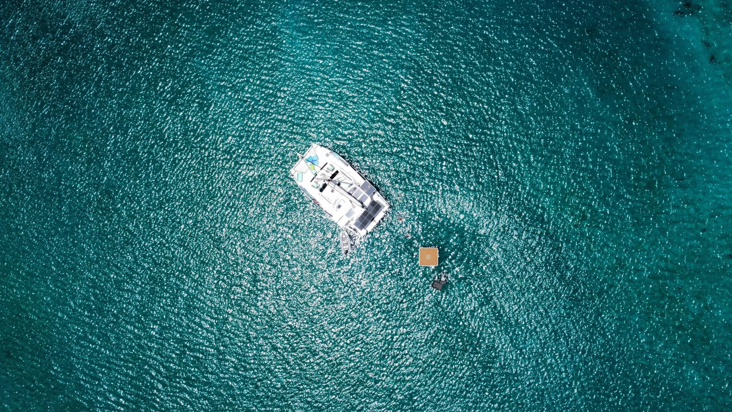 Aerial view of a white yacht anchored in turquoise water, with a small dinghy nearby and a square platform with a person sitting underneath.