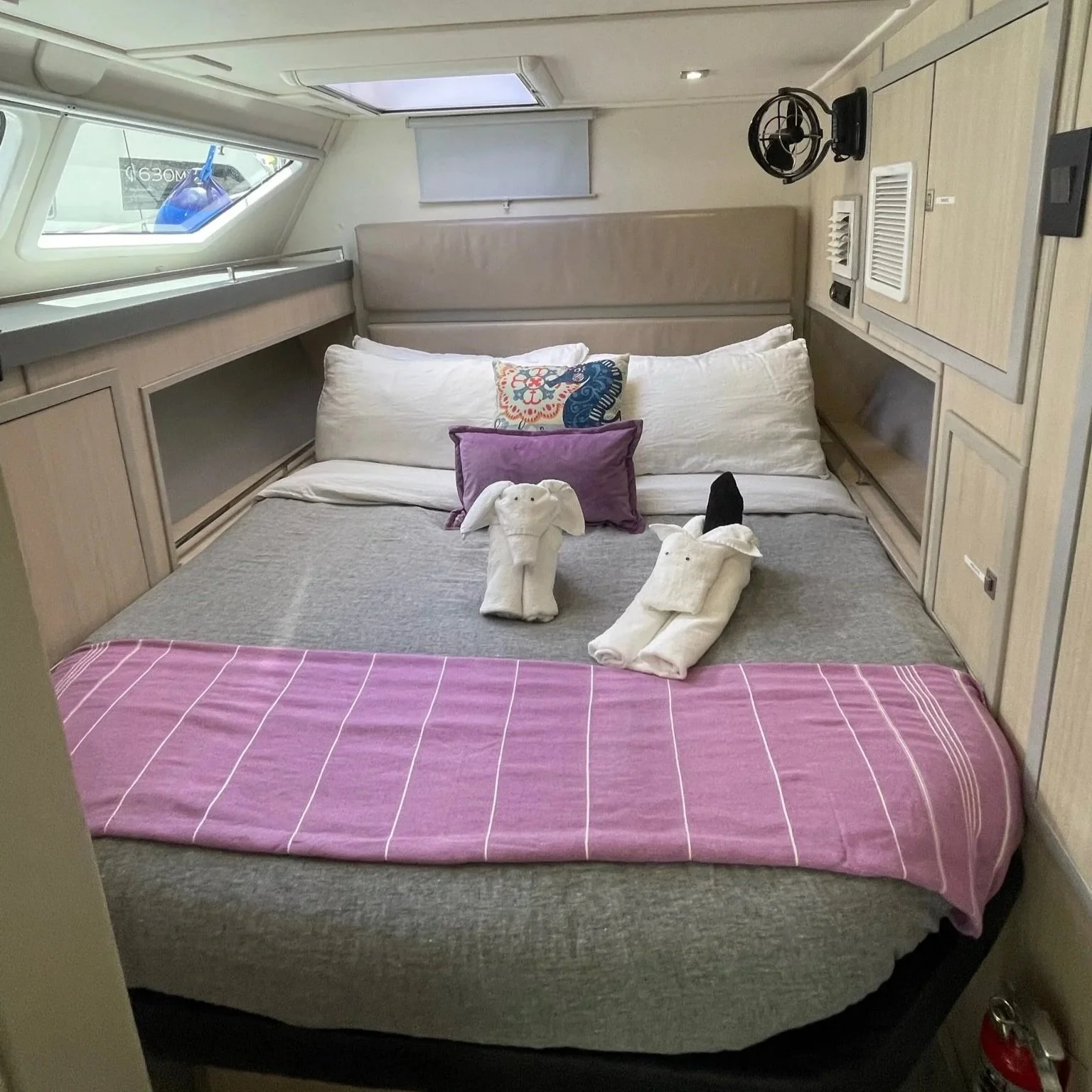 A bedroom on a boat or yacht with a bed covered in a pink and gray-striped bedspread, decorated with white pillows, and two towel animals resembling rabbits placed on the bed. There are built-in storage compartments and a small fan on the right side, with windows, one on the ceiling and another on the side, allowing natural light to enter.