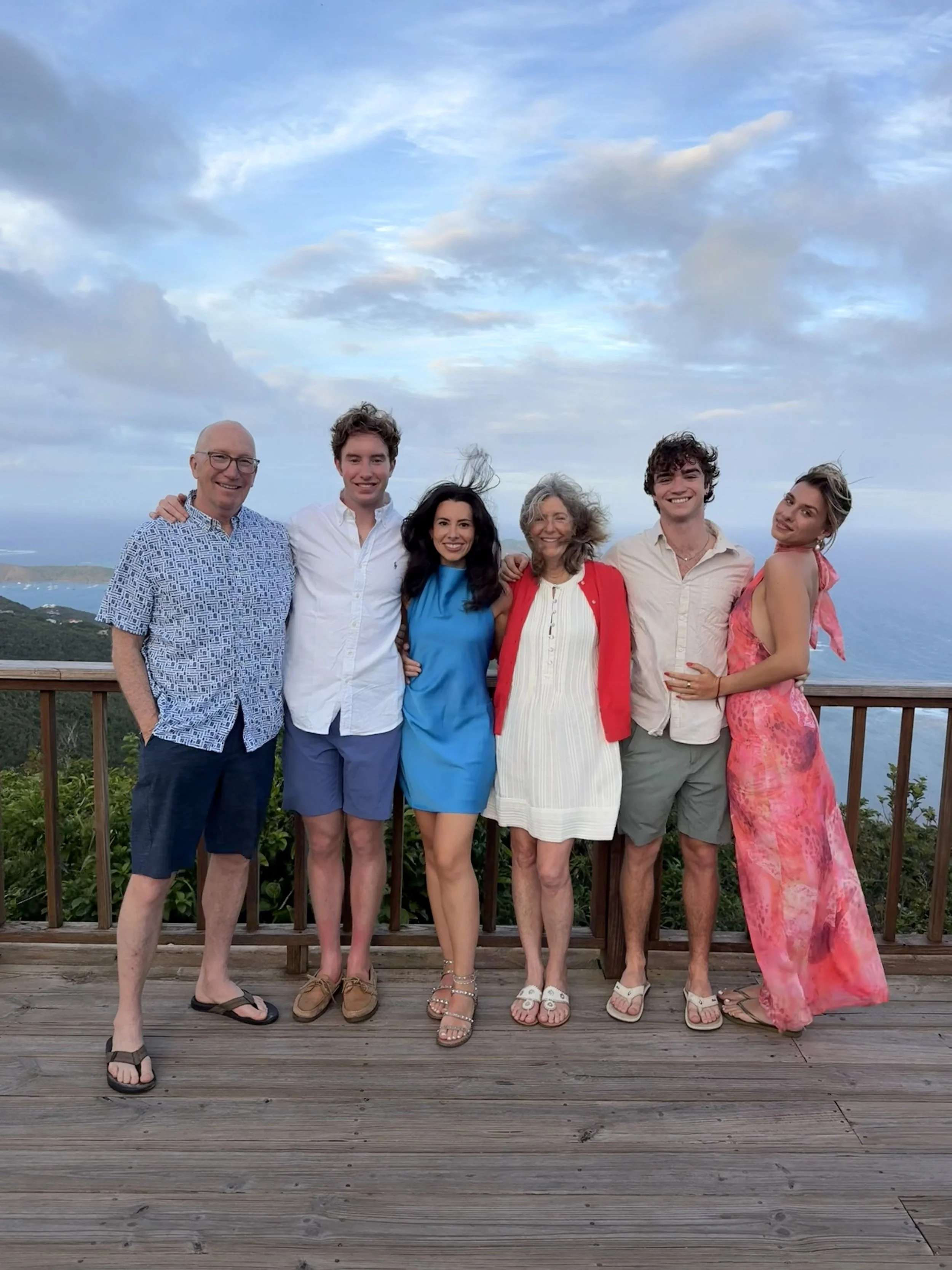 A group of seven people standing on a wooden deck outdoors, with a scenic view of the sky and distant landscape in the background. They are smiling and have their arms around each other.