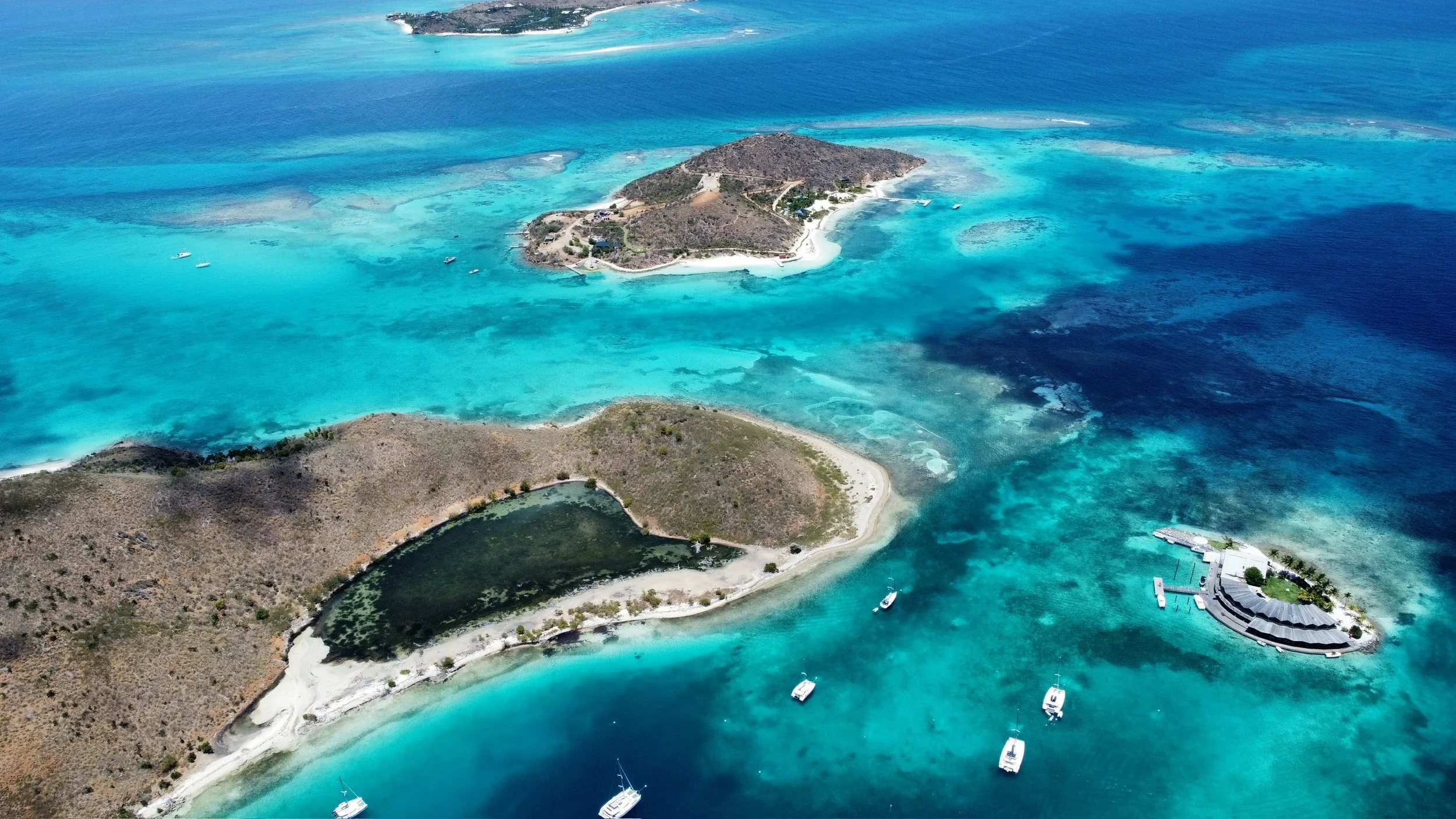 Aerial view of a tropical island with surrounding turquoise waters, boats anchored nearby, and a small marina with a curved pier on the right.