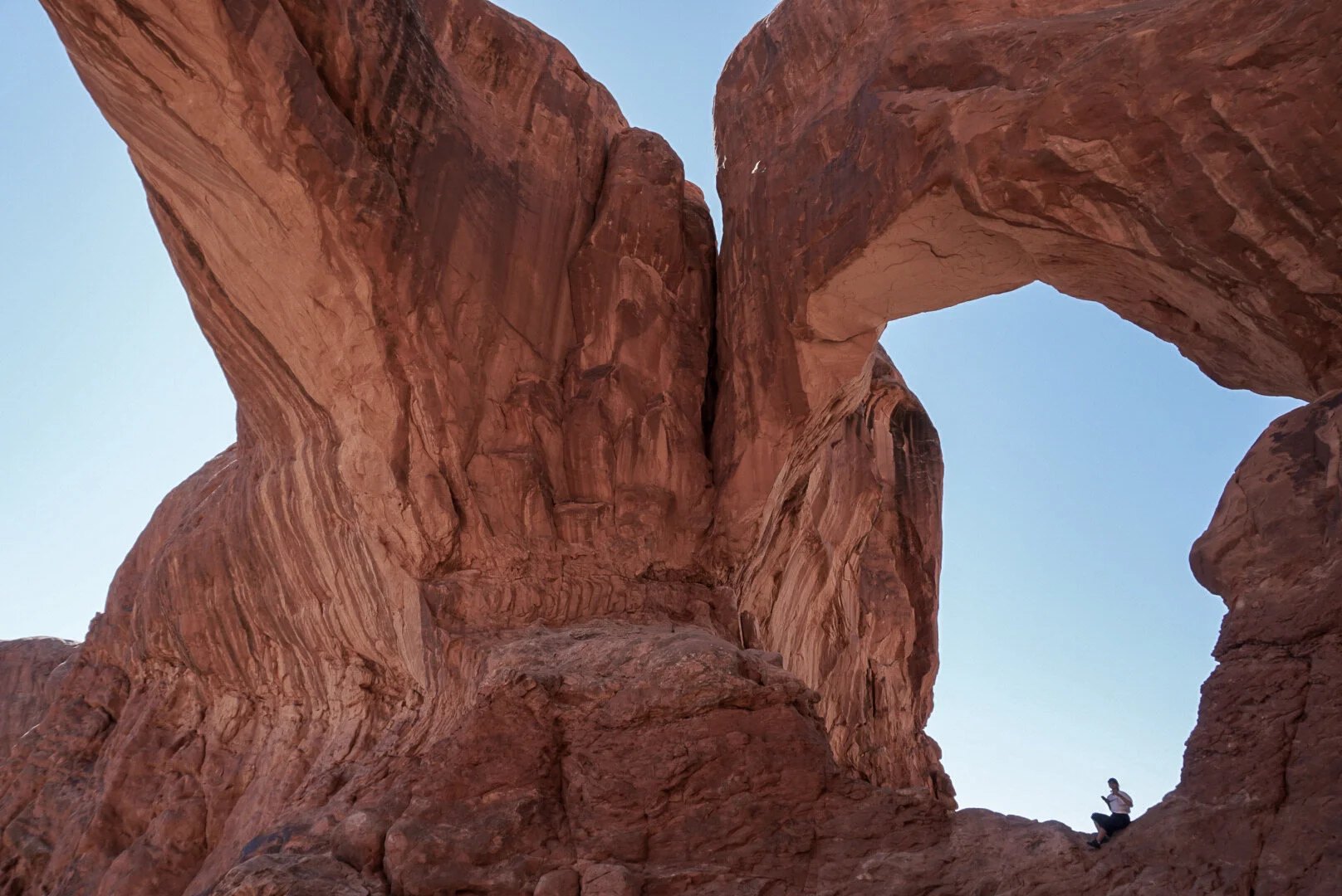 A person sitting on a large rock formation in front of a massive, natural rock arch in a desert landscape with red sandstone cliffs and a clear blue sky.