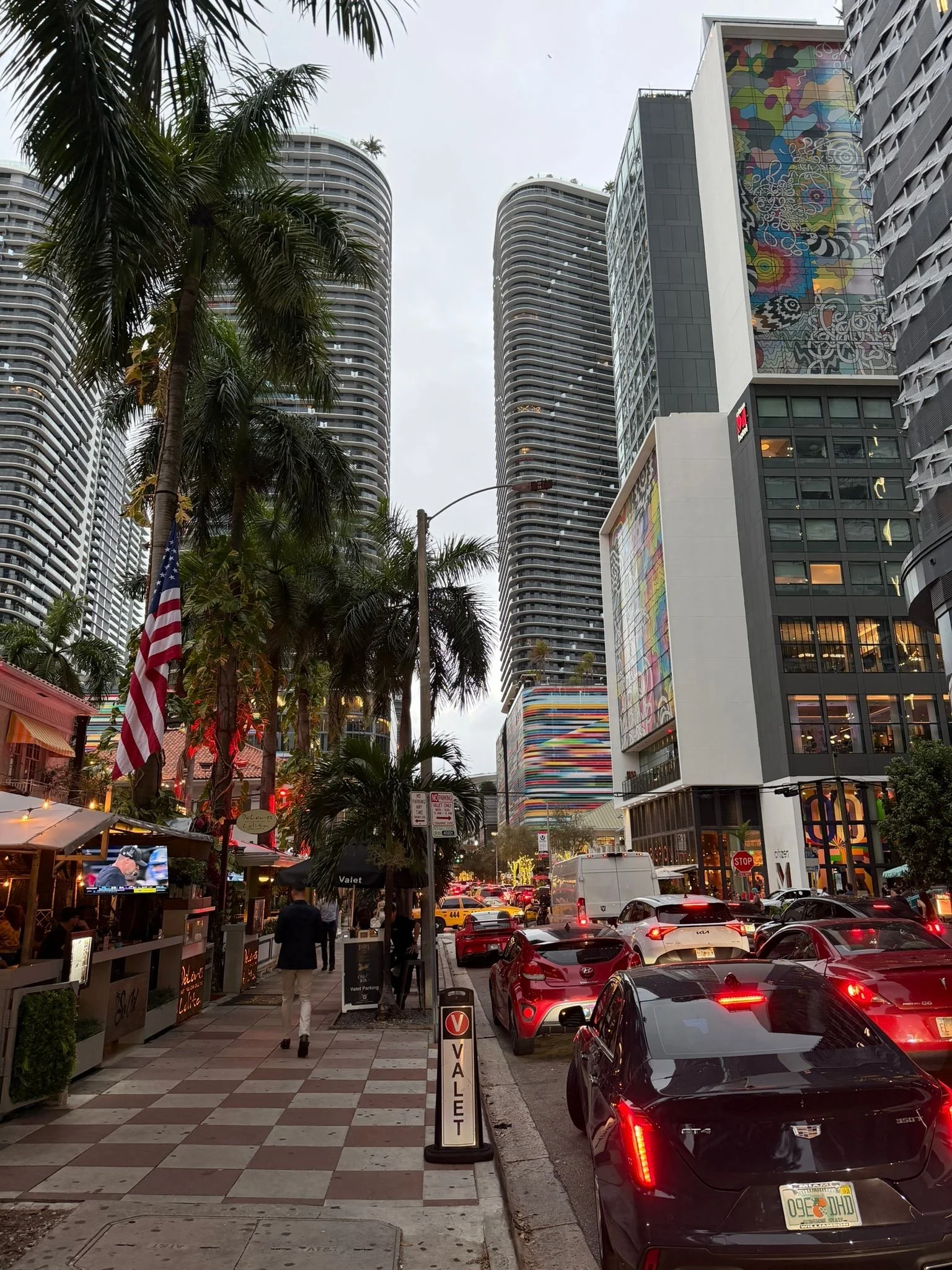 Bygate i Miami med høye, moderne skyskrapere, palmer, fargerike fasader og tett trafikk ved restauranter og butikker i kveldslys.