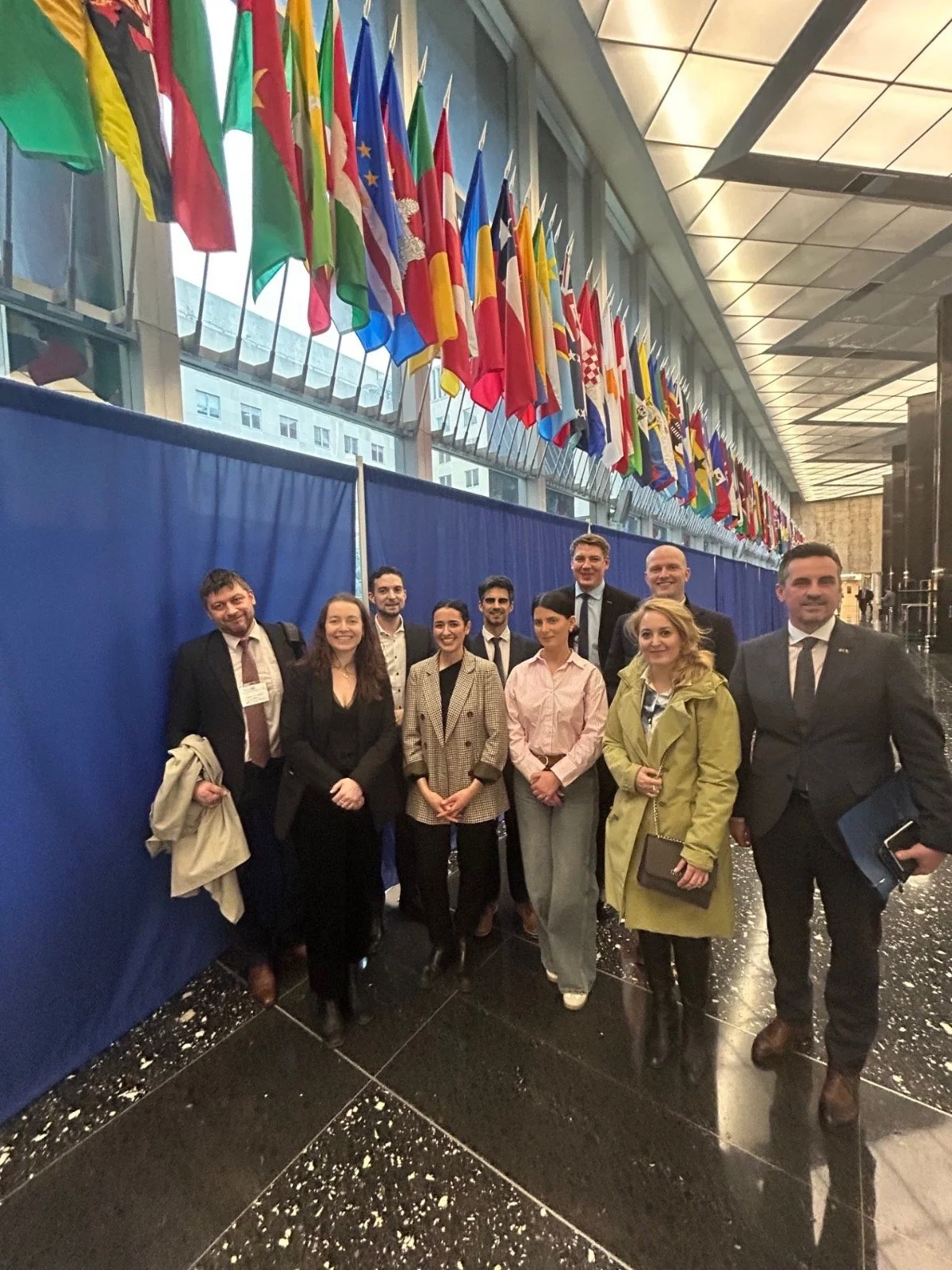 En gruppe deltakere står samlet i en hall dekorert med et langt galleri av internasjonale flagg, med blå skillevegger og et moderne glasstak i bakgrunnen