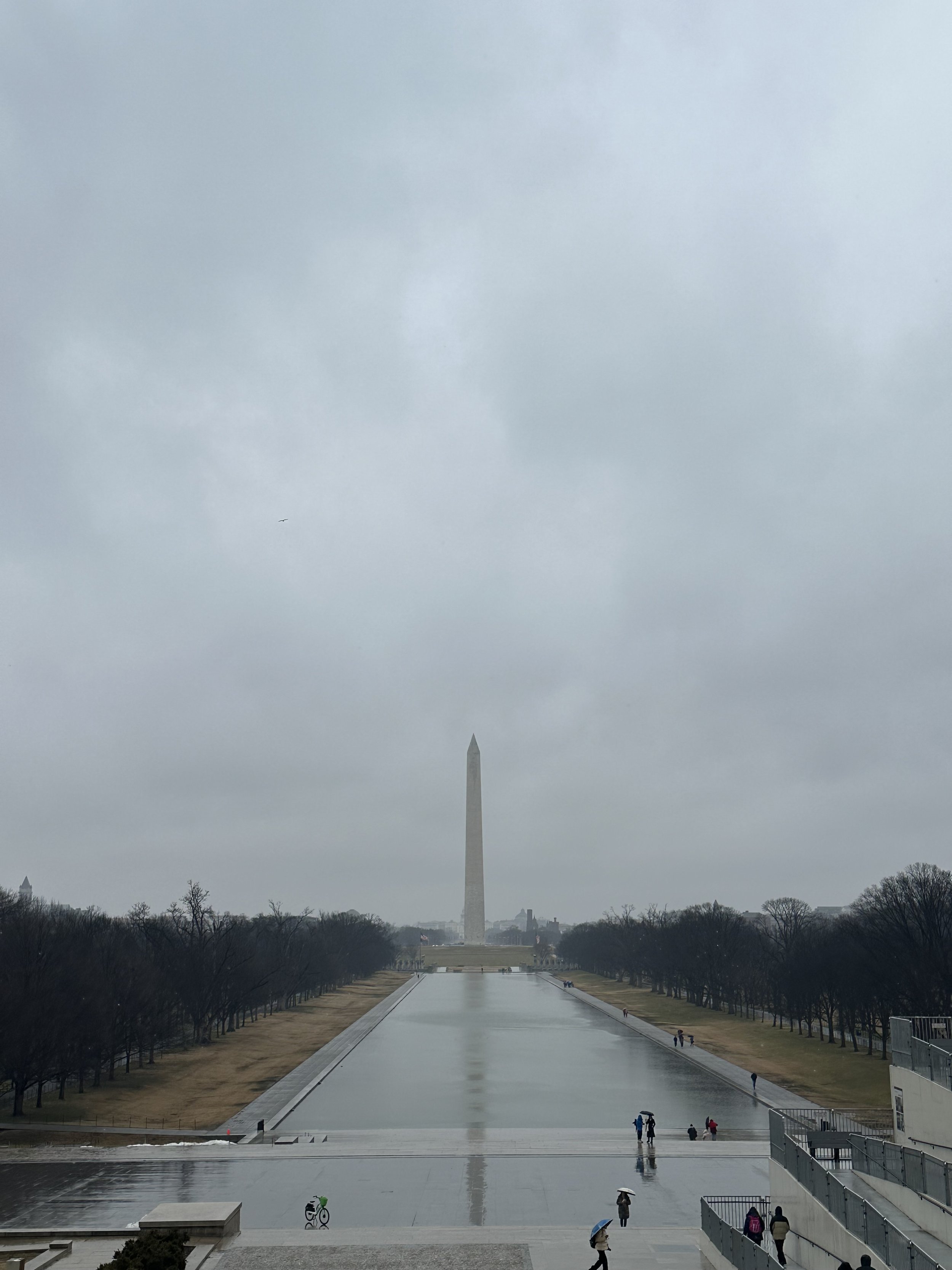 Utsikt over Reflecting Pool i Washington D.C. på en grå og regntung dag, med Washington Monument i det fjerne og noen få mennesker som går langs kantene av vannspeilet.