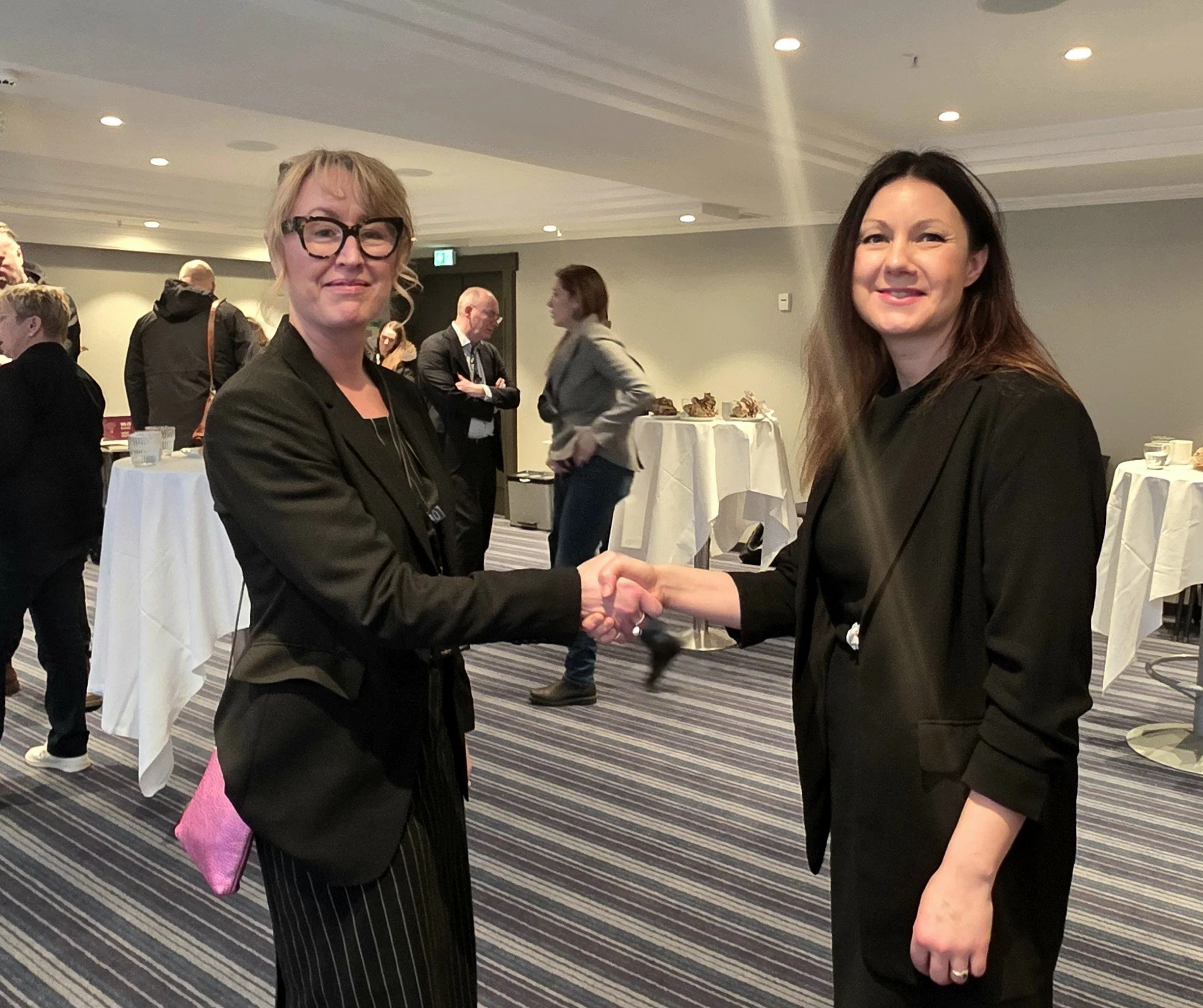 To personer i mørke klær håndhilser i et konferanserom med ståbord og flere andre deltakere i bakgrunnen. Rommet har stripet teppe, dempet belysning og et uformelt minglemiljø.