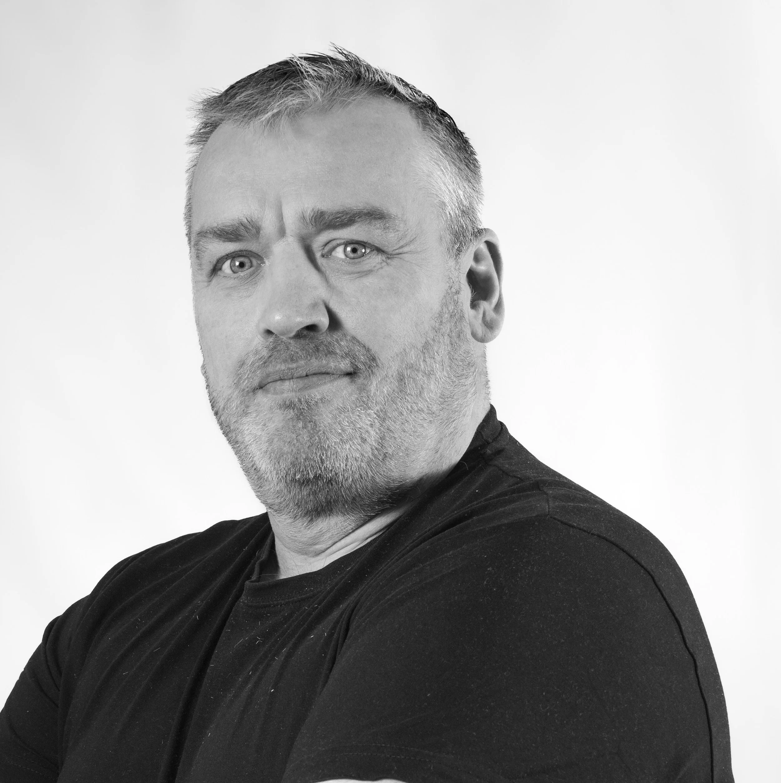 Sort-hvitt portrettfoto av en person fotografert fra brystet og opp, med armene krysset. Ansiktet er delvis tildekket av en grå blokk. Lys bakgrunn