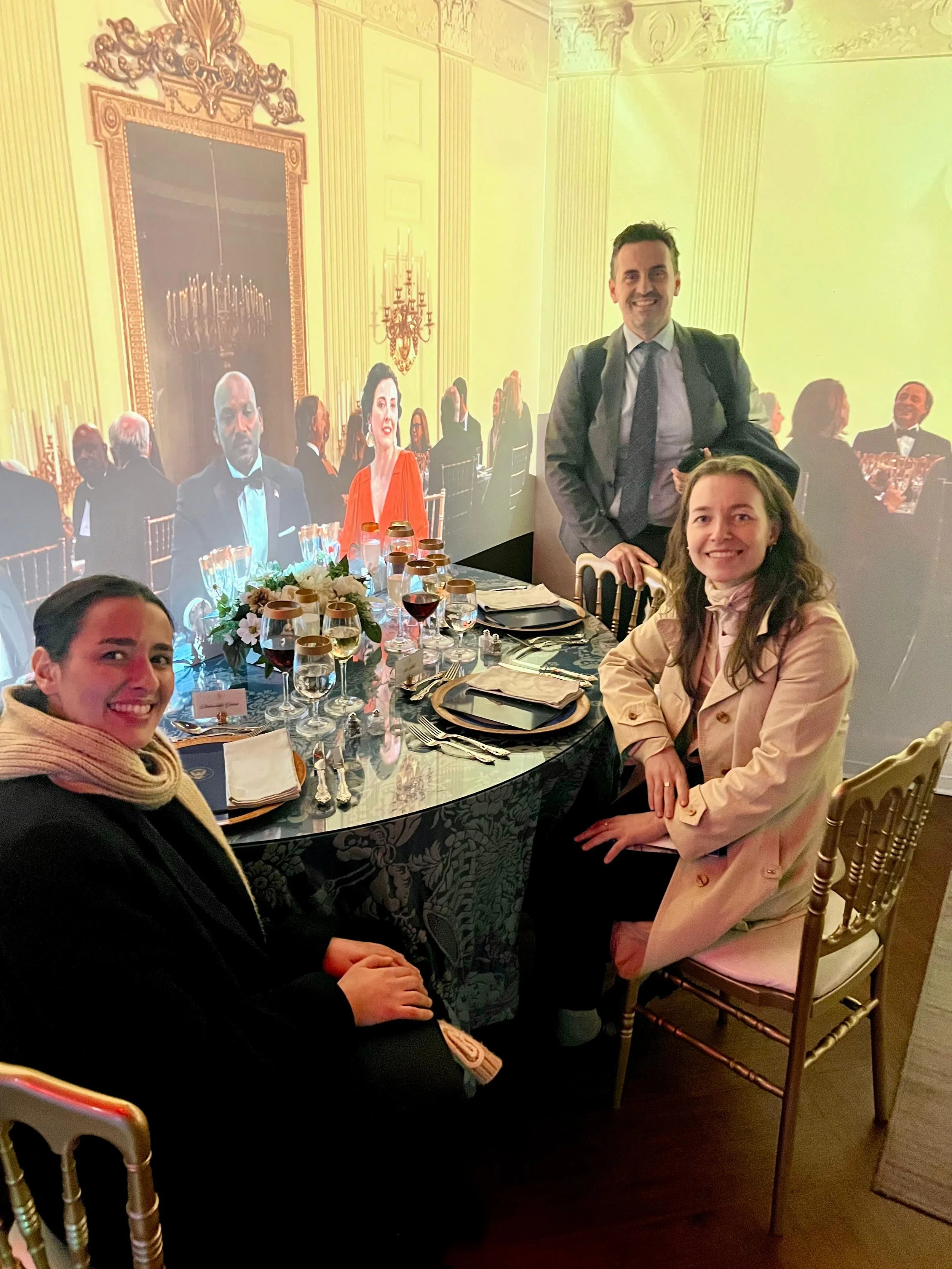 En gruppe personer sitter og står rundt et elegant, festdekket bord i et rom med store projiserte bilder av en formell bankettsal i bakgrunnen.