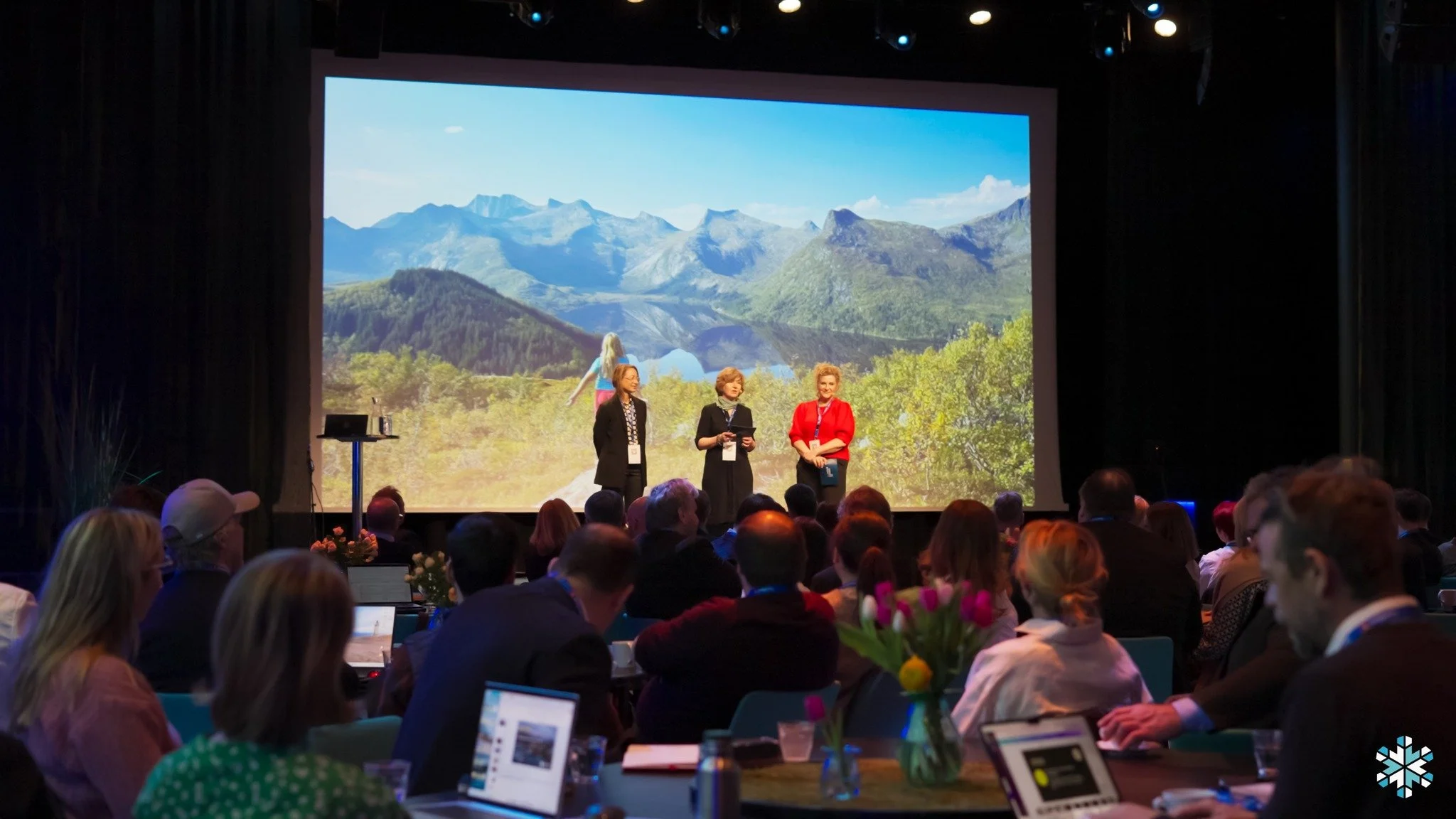 Konferansesal der tre personer står på scenen foran et stort lerret som viser fjellandskap, mens publikum sitter ved bord med skjermer og notater.