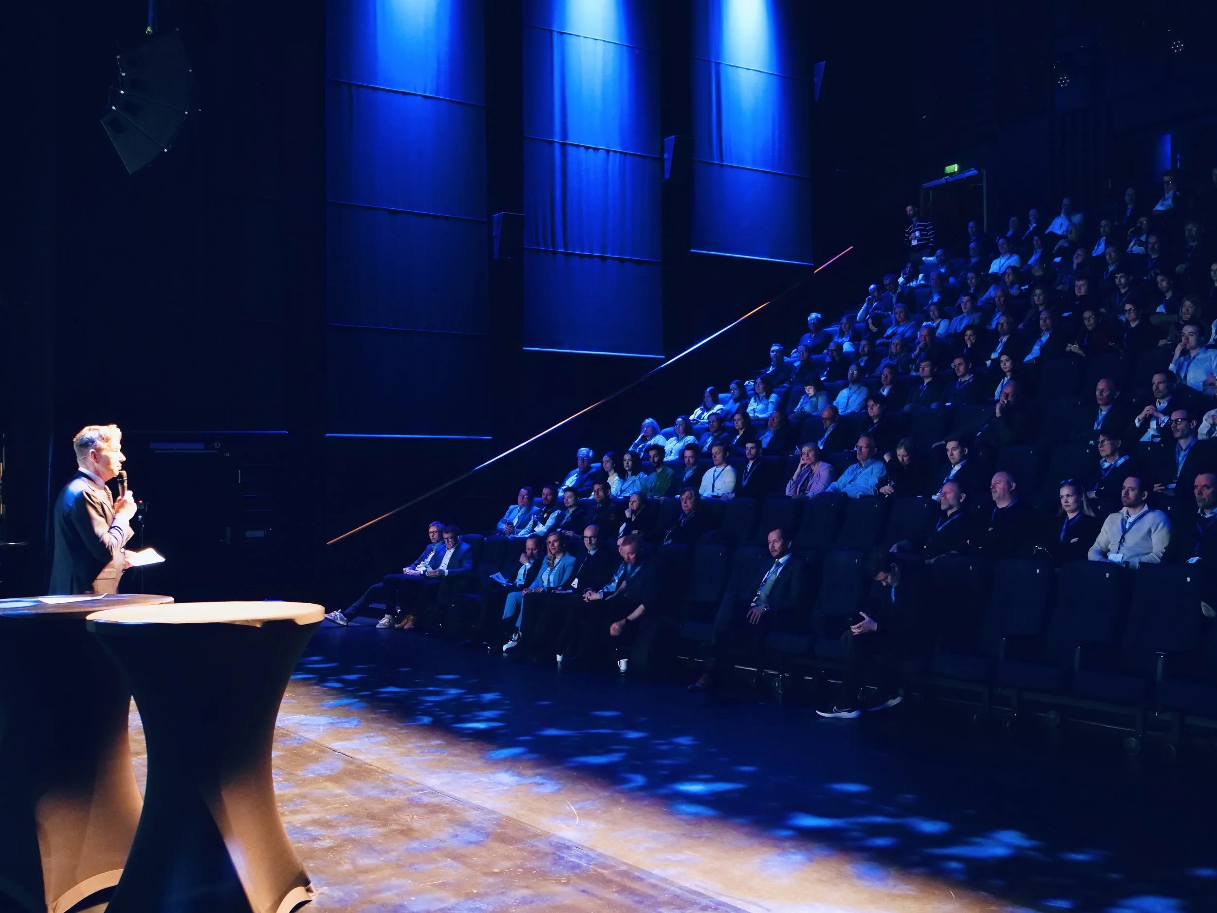 Foredragsholder står på scenen i et mørklagt auditorium, mens et stort publikum sitter i amfi belyst av blått scenelys