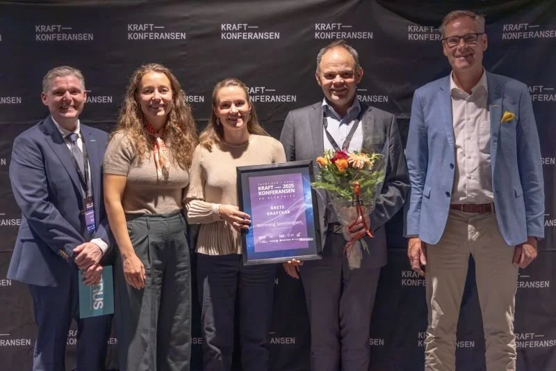 Fem personer står foran en bakvegg med logoen til Kraftkonferansen; én holder en innrammet pris og en blomsterbukett.