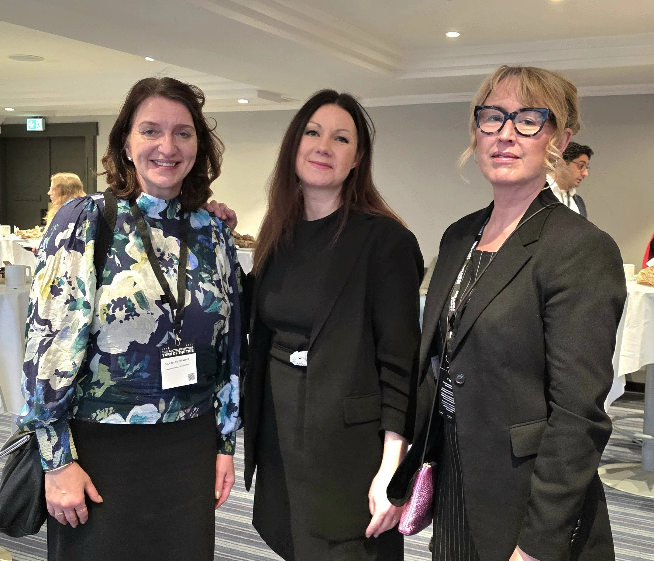 Tre personer står sammen i et konferanserom, kledd i profesjonelle antrekk. De smiler mot kameraet, og bak dem står høye bord med hvite duker og andre deltakere som mingler i rommet.