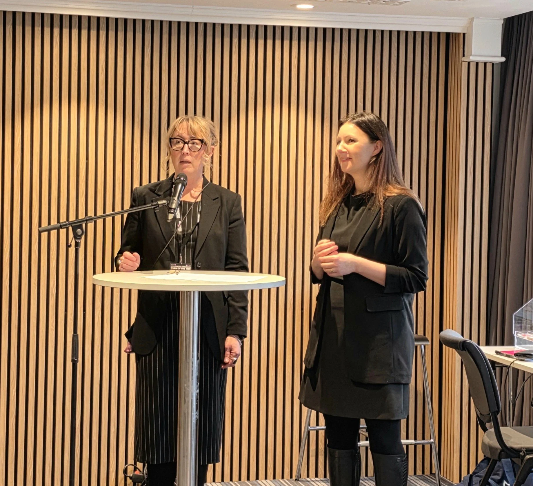 To personer står ved et høyt rundt bord foran en mikrofon i et rom med vertikale trefiner‑paneler. Begge er kledd i mørke, profesjonelle antrekk og ser ut til å delta i en presentasjon eller samtale.