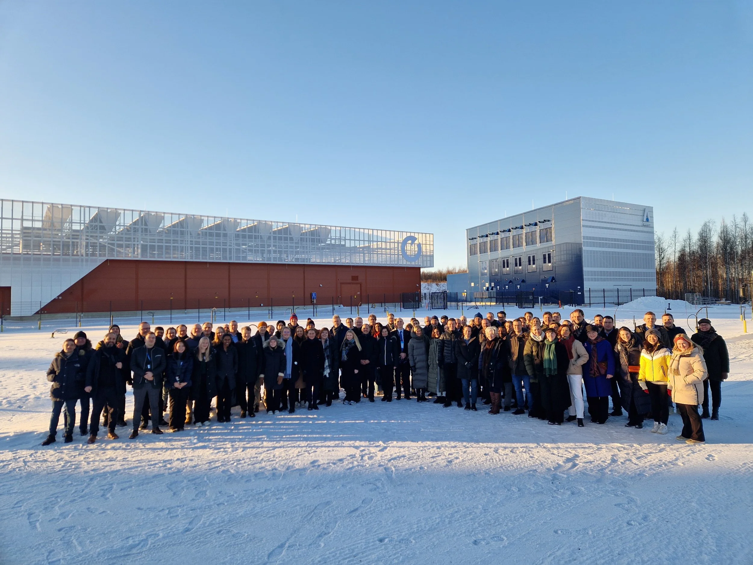 Stor gruppe mennesker står ute på et snødekt område foran industribygg i sollys.