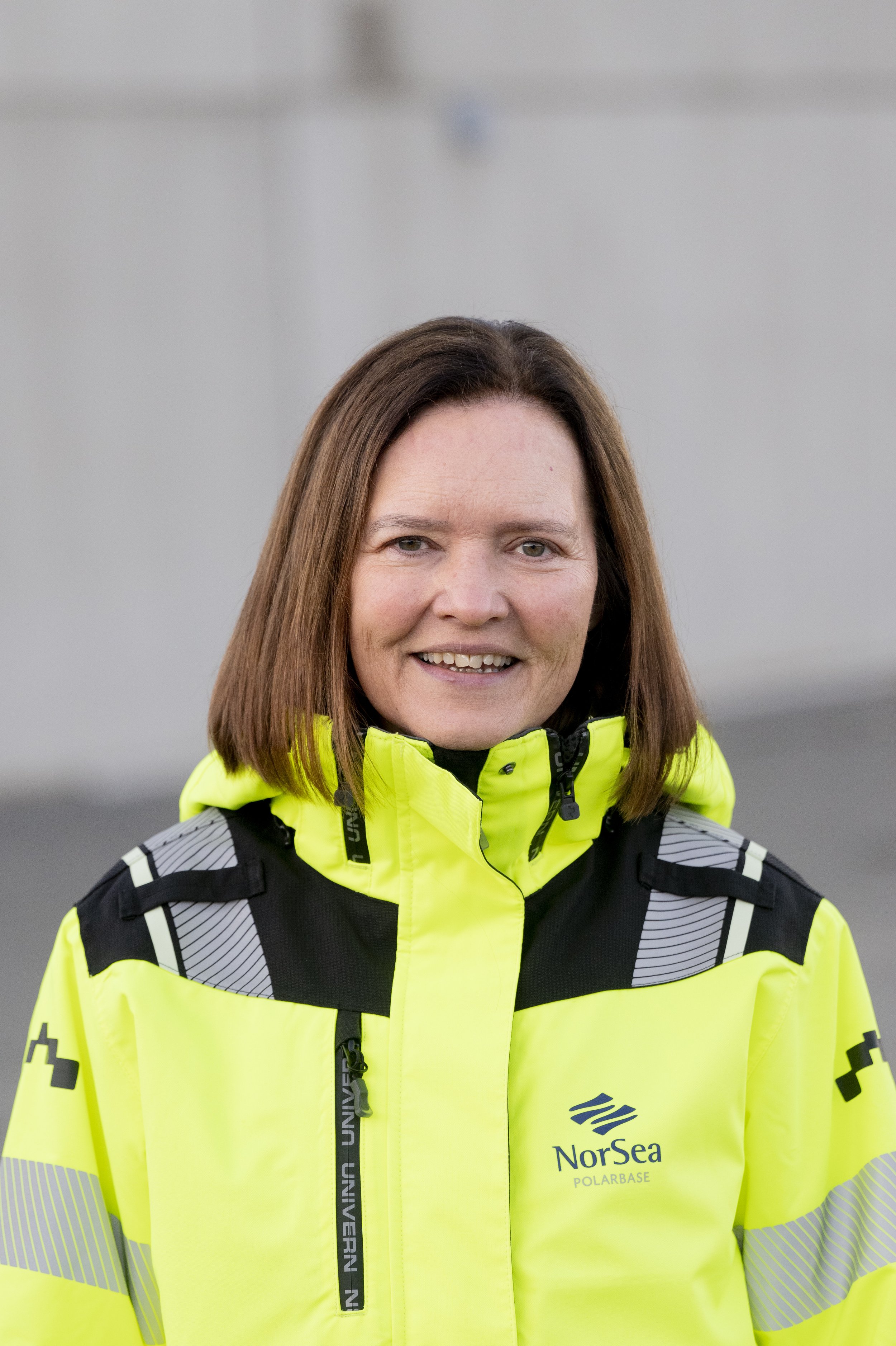 Person iført en gul høysynlighetsjakke med svarte og grå detaljer, merket med NorSea Polarbase‑logoen, står utendørs foran en nøytral, grå bakgrunn.