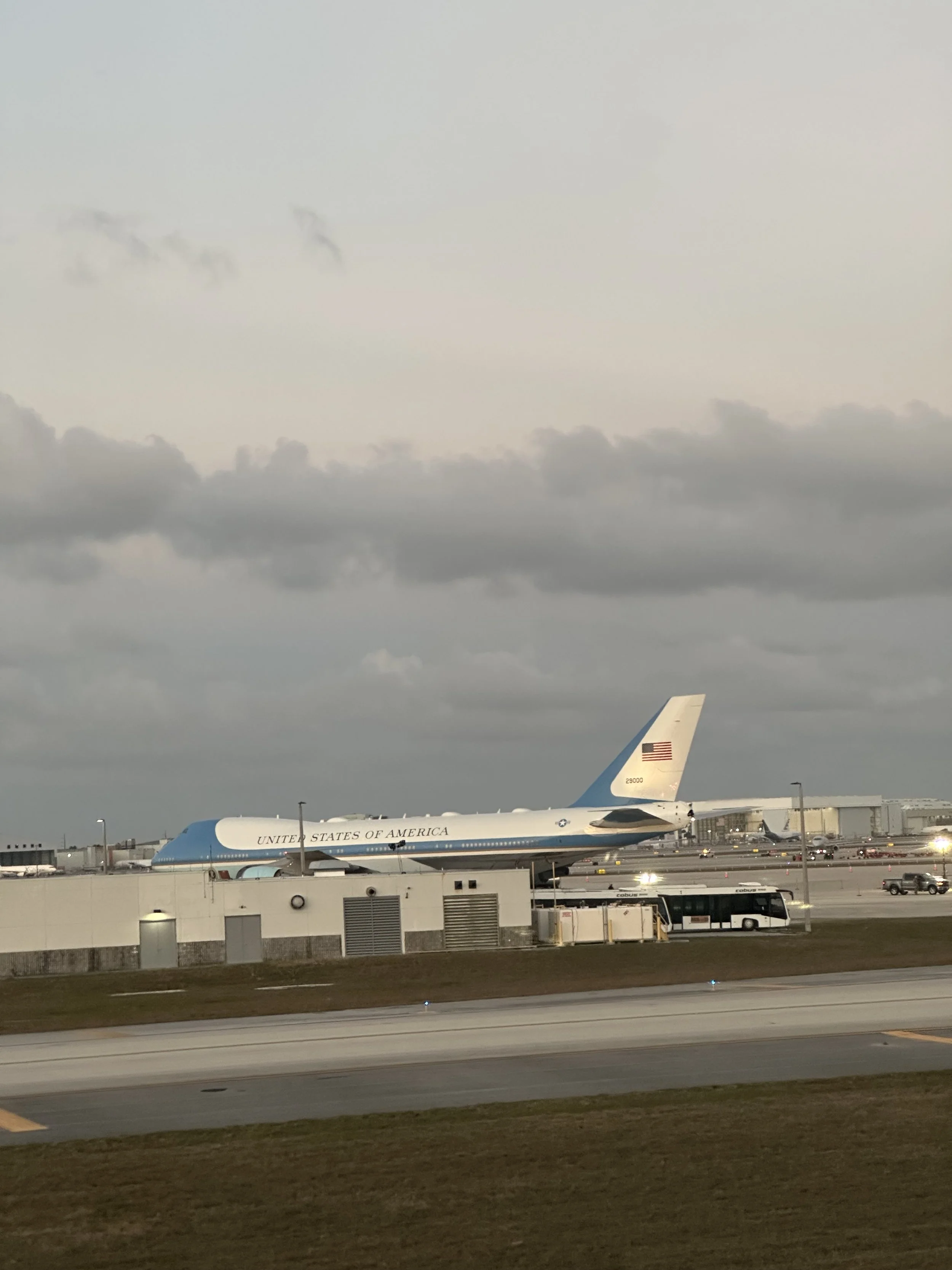 Flyet som brukes av USAs president står parkert på rullebanen ved en flyplass i kveldslys, omgitt av lave bygninger og kjøretøy, med skyet himmel i bakgrunnen