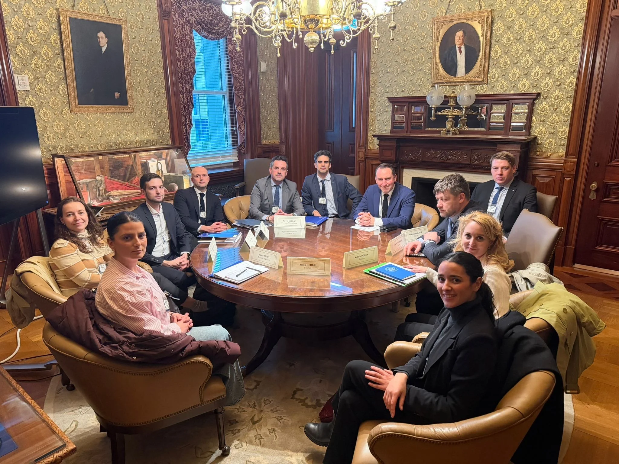 Møte rundt et ovalt konferansebord i et klassisk, historisk innredet rom, der flere deltakere sitter samlet til faglig diskusjon. Rommet har trepanel, utsmykkede tapeter, lysekrone og portretter på veggene