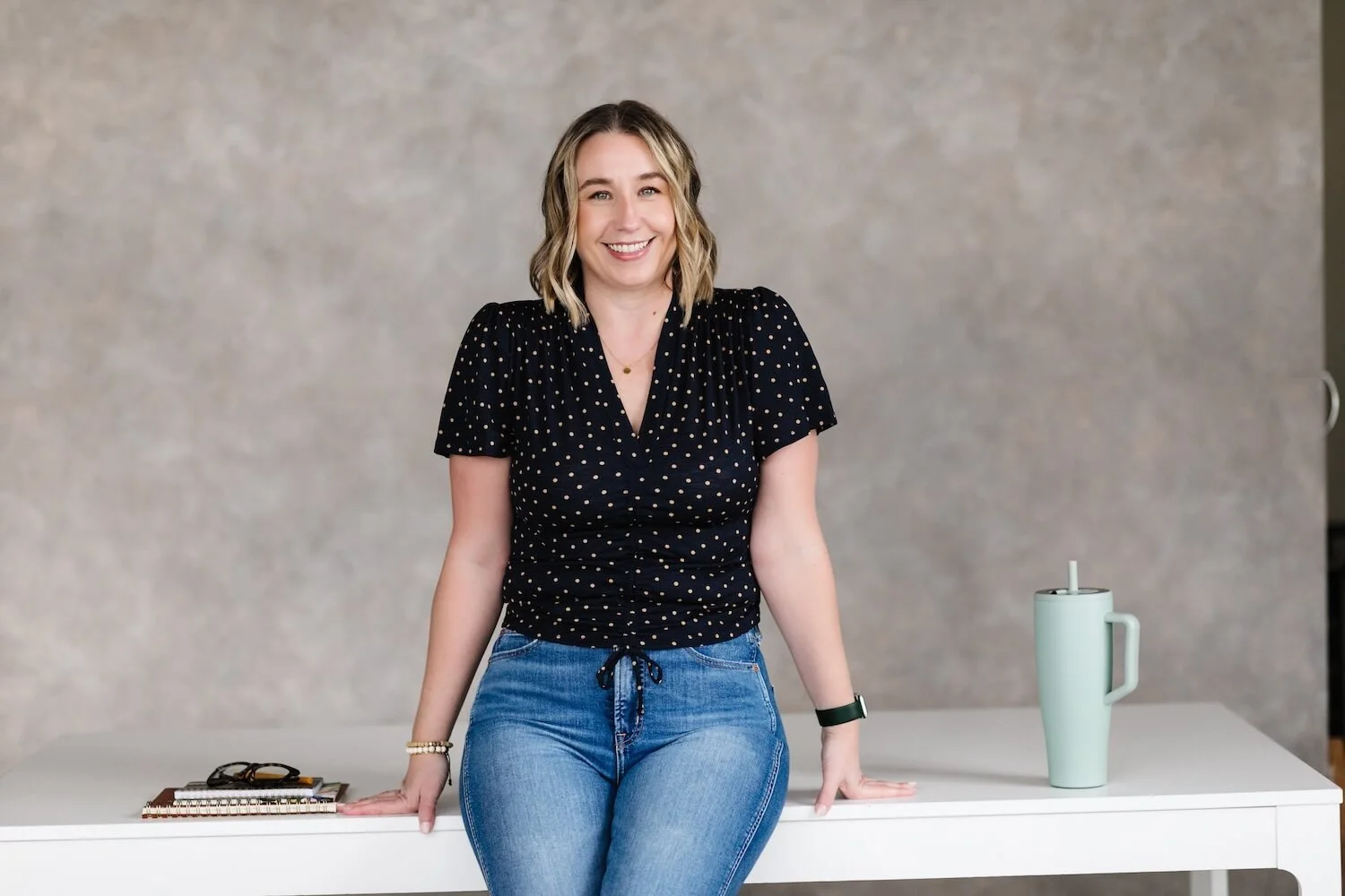 A woman sitting on a white table, smiling, with a gray background, a notebook, glasses, and a light green tumbler with a lid and straw nearby.