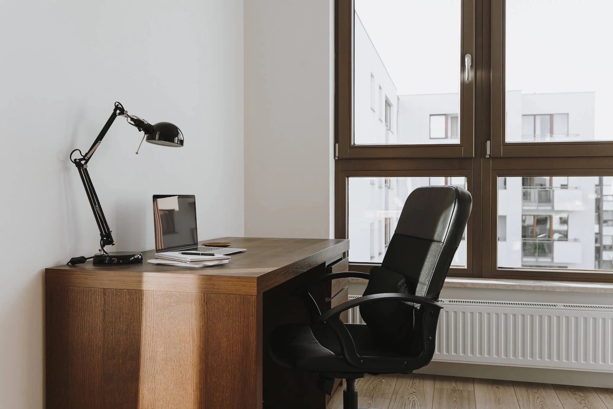 A minimalist office with a wooden desk, black ergonomic chair, desk lamp, laptop, notebooks, and pens near large windows with a view of neighboring apartment buildings.
