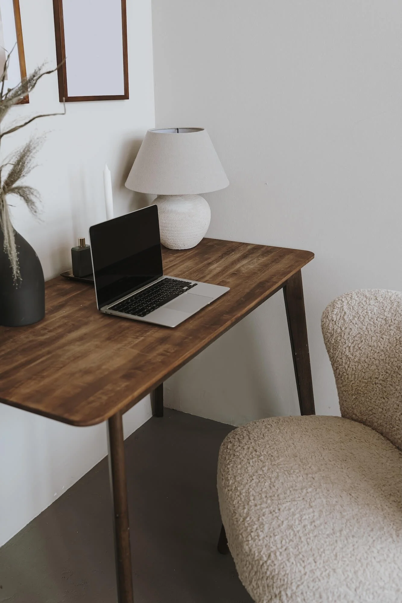 A wooden desk with a black laptop, a white textured lamp, a black vase with dried plants, and a white candlestick is positioned next to a beige plush armchair in a minimalist room with white walls.