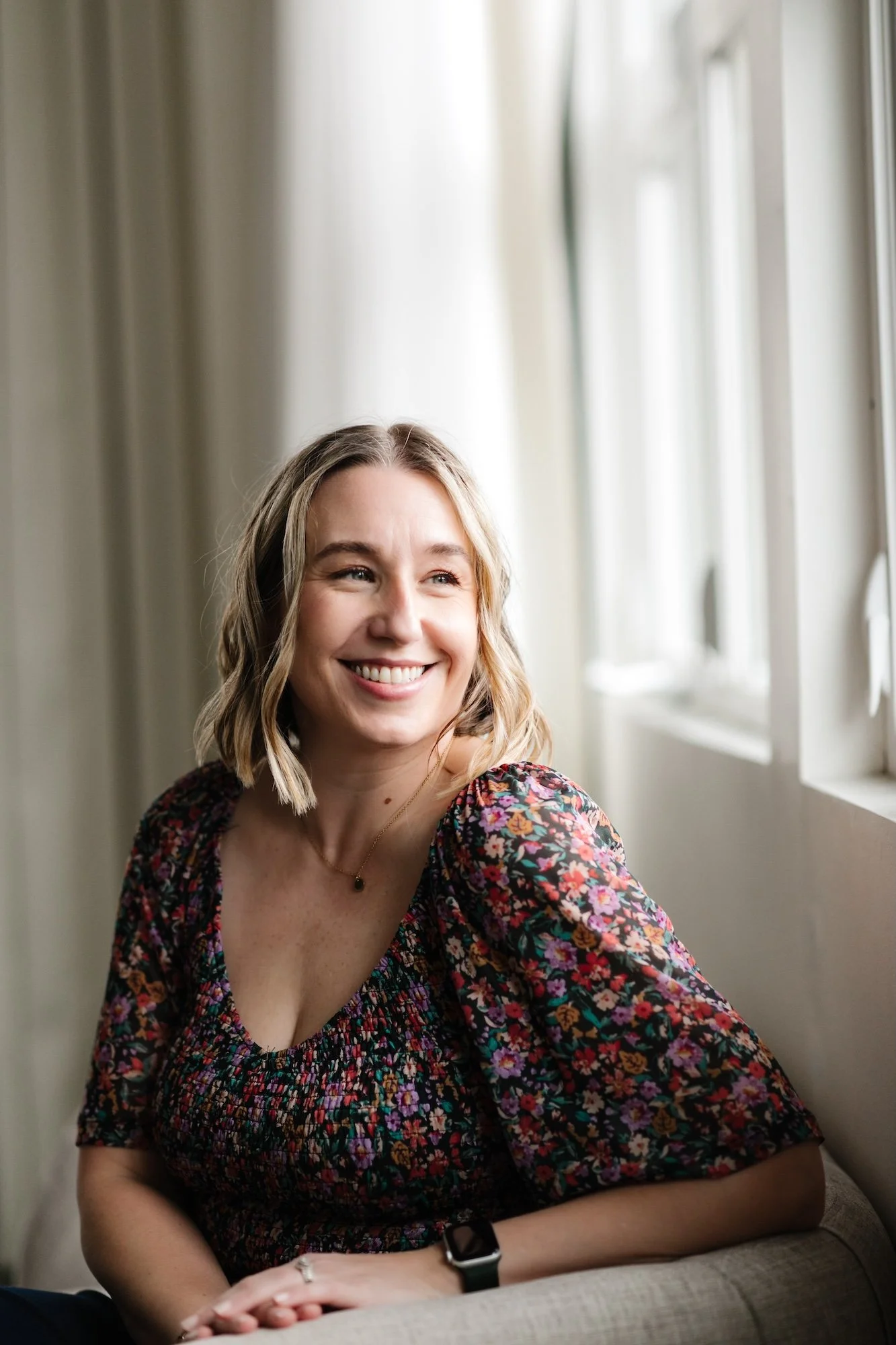Smiling woman with short blonde hair sitting by a window in a floral top, looking out and enjoying the sunlight.