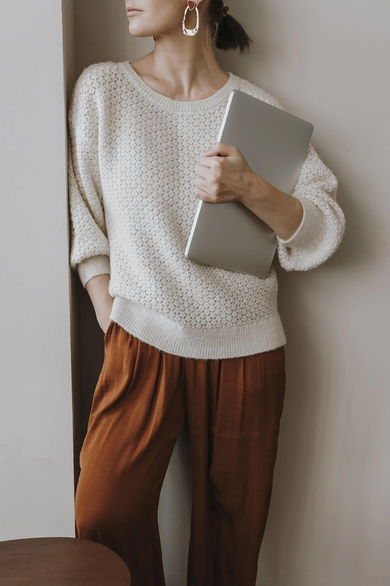 A woman standing against a wall, holding a closed laptop, wearing a cream crochet sweater, brown pants, and large earrings.