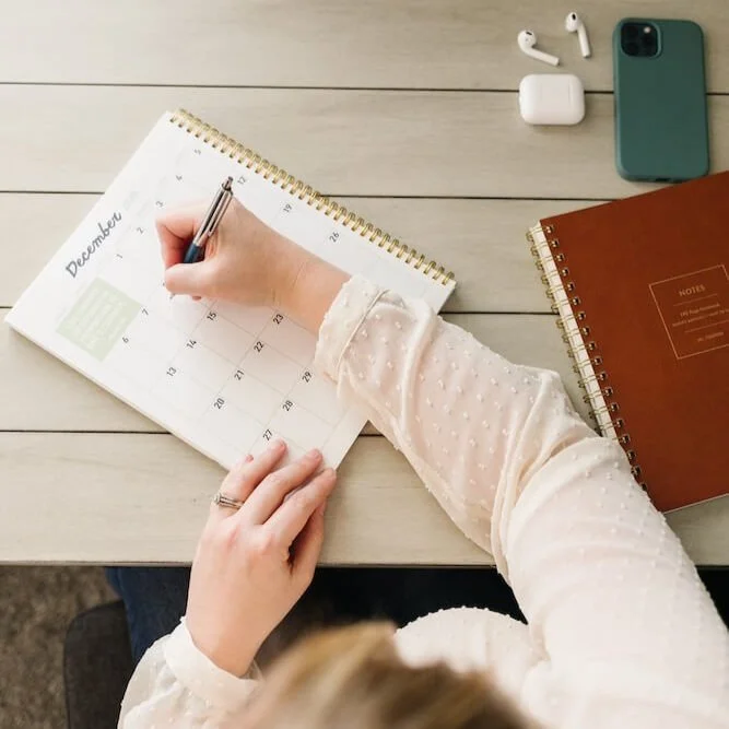 A person writing in a December calendar with a black pen, on a light-colored desk. Around the calendar are wireless earbuds, a smartphone, and notebooks.
