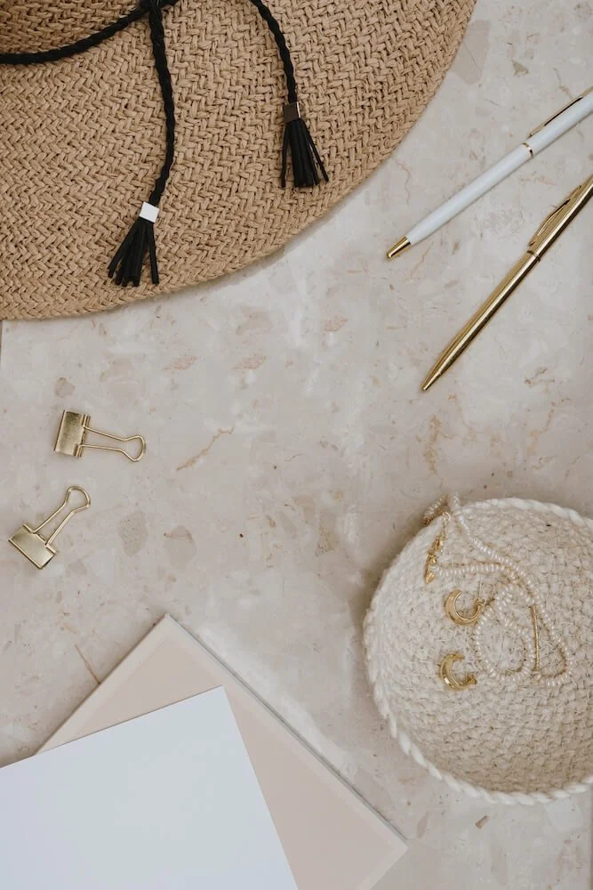 Flat lay of beige woven handbag, white and gold pens, gold paper clips, white pearl jewelry, and a beige knitted round container with gold jewelry, on a light marble surface.