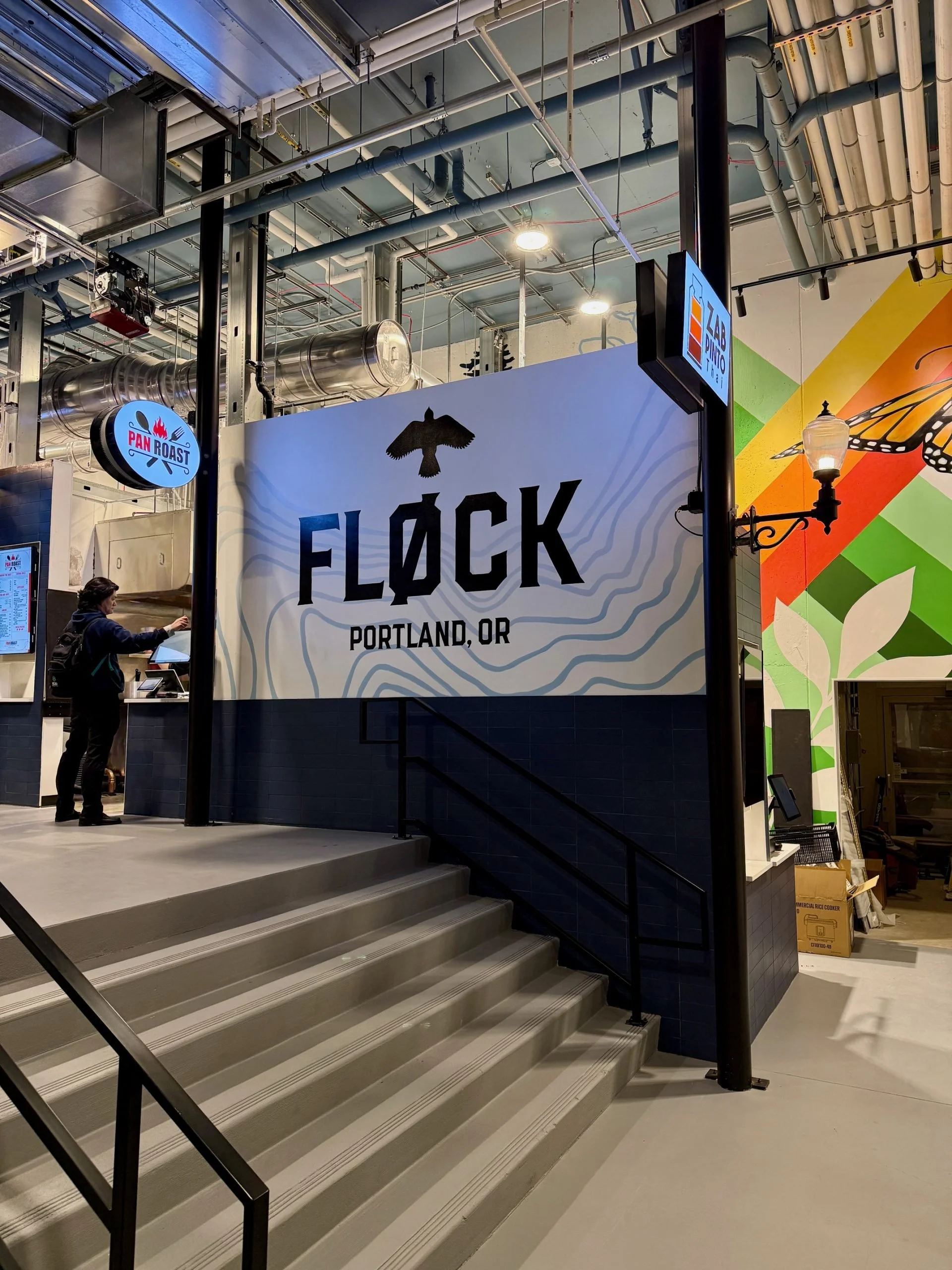Indoor view of Flock restaurant in Portland, Oregon, with a large sign featuring a black bird silhouette and the text 'FLꓘCK PORTLAND, OR.' The surrounding area includes stairs, a person using a touchscreen ordering kiosk, and colorful wall murals.
