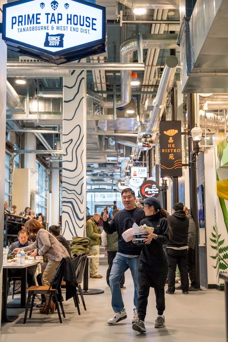 People dining and walking inside a food court or market. Signs for Prime Tap House and HK Bistro are visible. The area has modern industrial decor with exposed pipes and patterned columns.