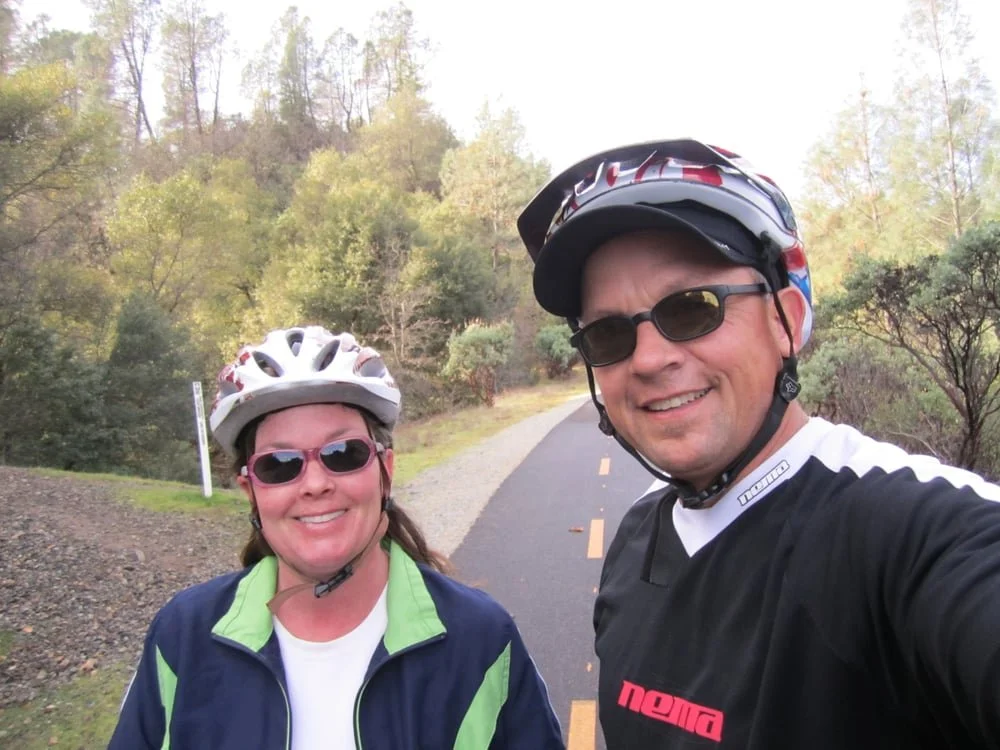 A man and woman taking a selfie on a biking trail surrounded by trees, both wearing helmets and sunglasses.