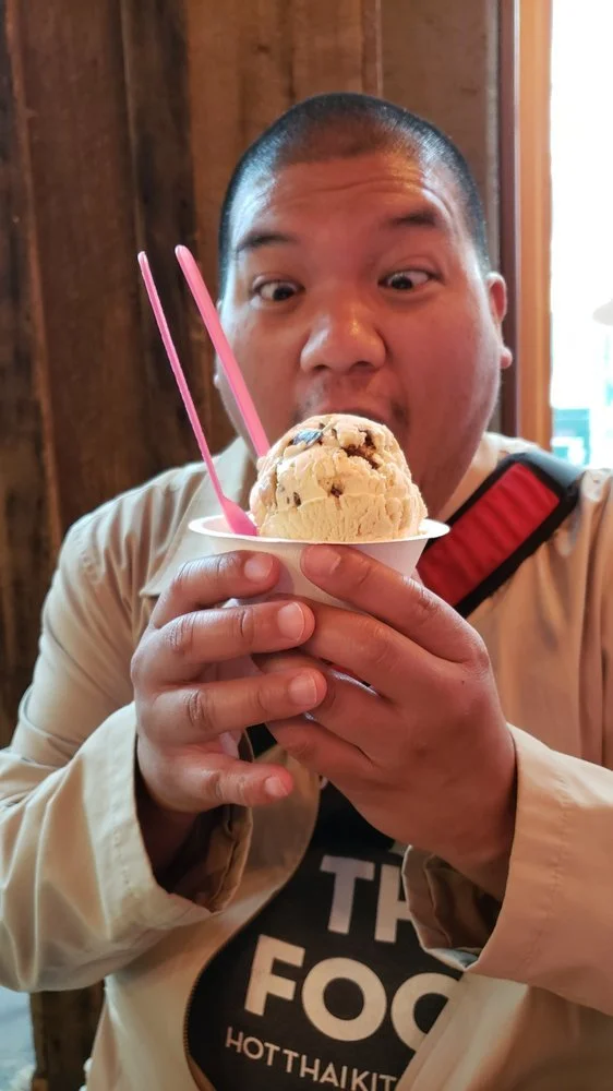 A man with short hair holding a cup of cookie ice cream with pink plastic spoons, about to eat it, indoors with wooden walls in the background.