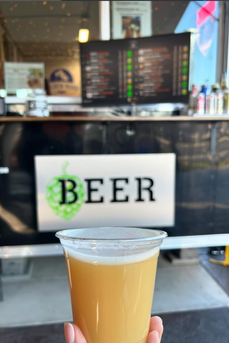 A hand holding a plastic cup filled with a cloudy, light yellow beer in front of a sign that says "BEER" with a green hop illustration, at a beer stand or bar.