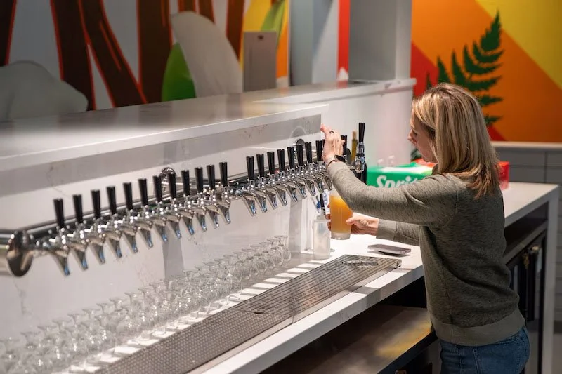 A woman in a gray sweater pours beer from a tap at a counter with multiple taps, in a modern bar or brewery setting.