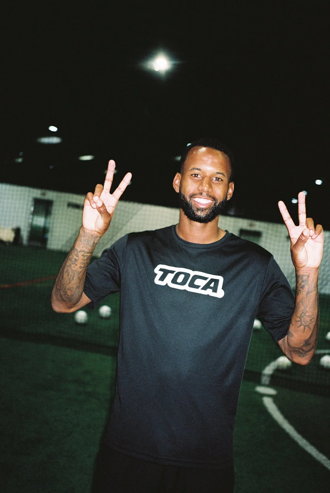 A man with a beard smiling, wearing a black T-shirt with the word TOCA, making peace signs with both hands inside an indoor sports facility with small white balls on the ground.
