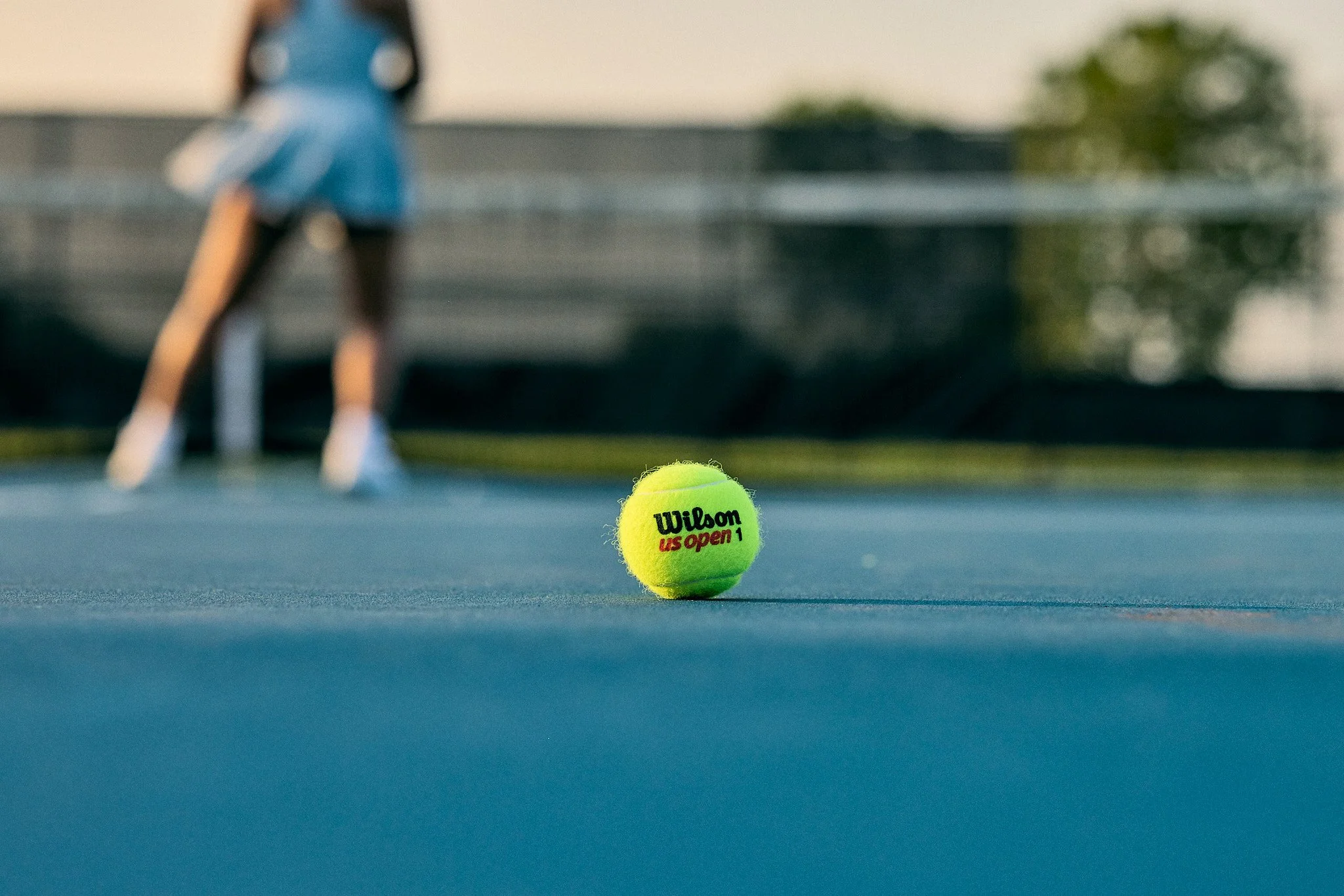 A close-up of a bright yellow tennis ball with the Wilson logo, placed on a blue tennis court with a person in a blue shirt and shorts in the background, slightly out of focus.