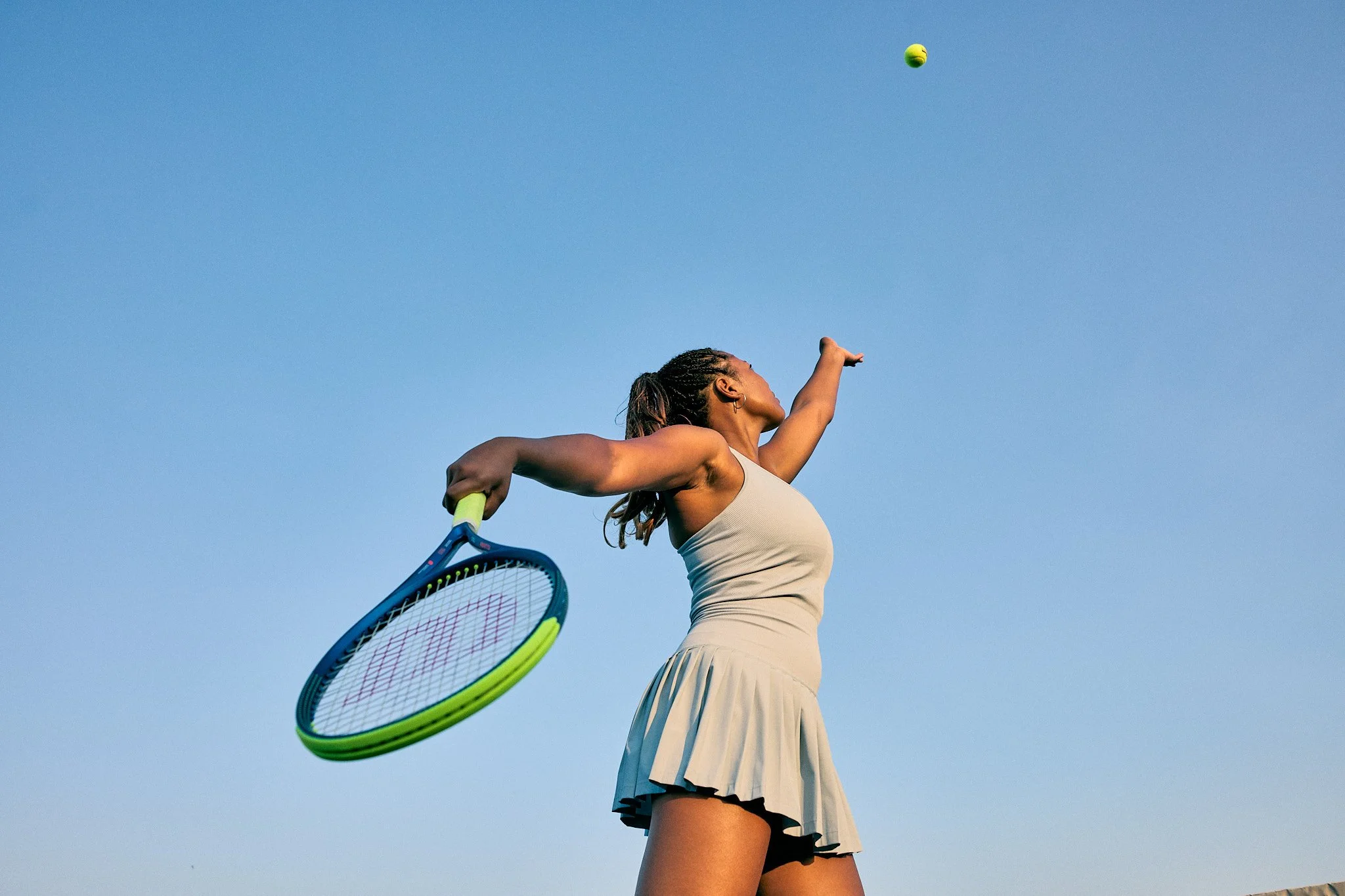 A woman in a beige tennis dress serves a tennis ball outdoors under a clear blue sky, holding a tennis racket in her right hand. Fitness, sports, commercial photography, Chicago based photographer. Wilson tennis