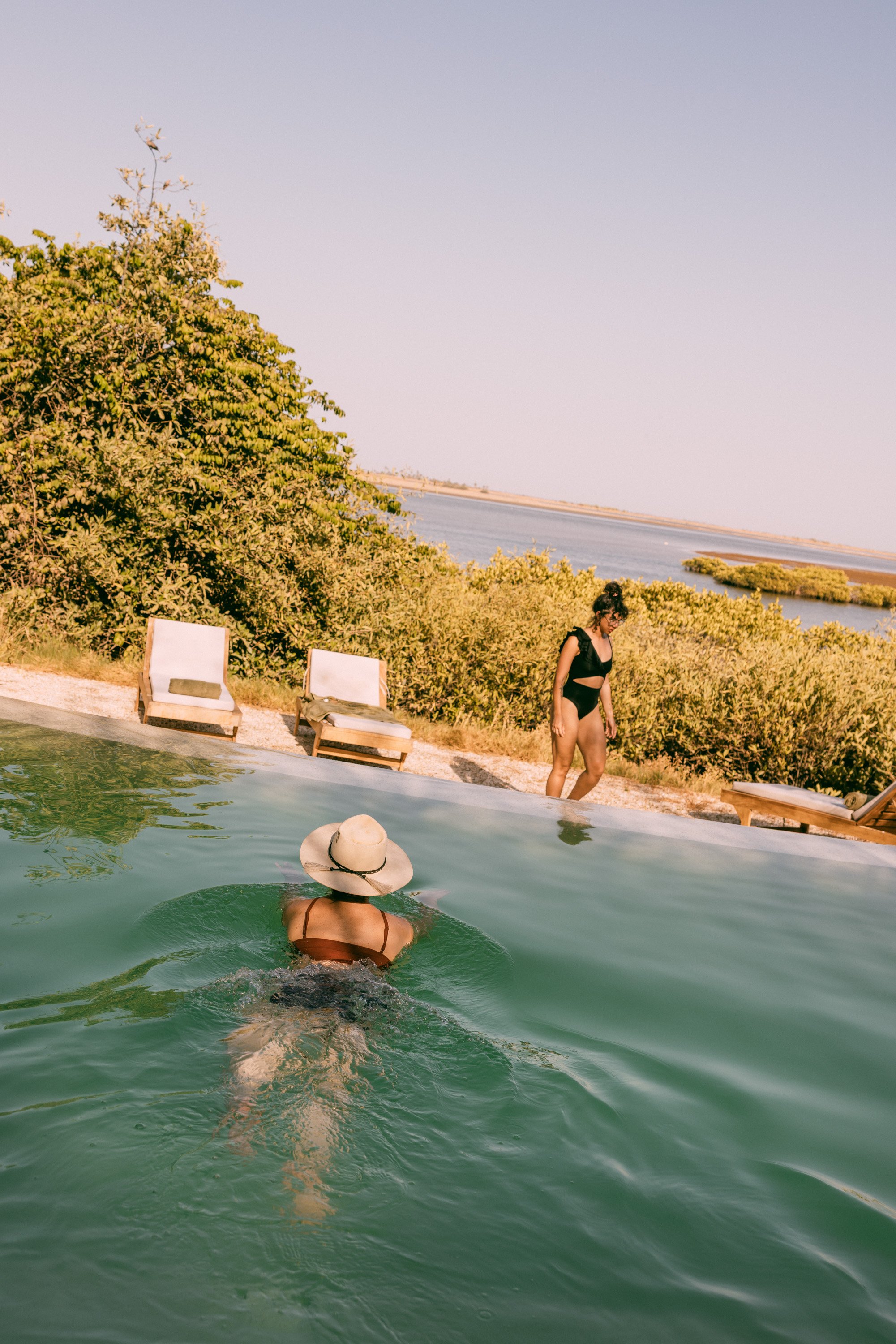 A woman in a black swimsuit with a wide-brimmed hat swimming in an outdoor pool near a sandy area with lounge chairs, overlooking a body of water and lush green bushes under a clear sky.