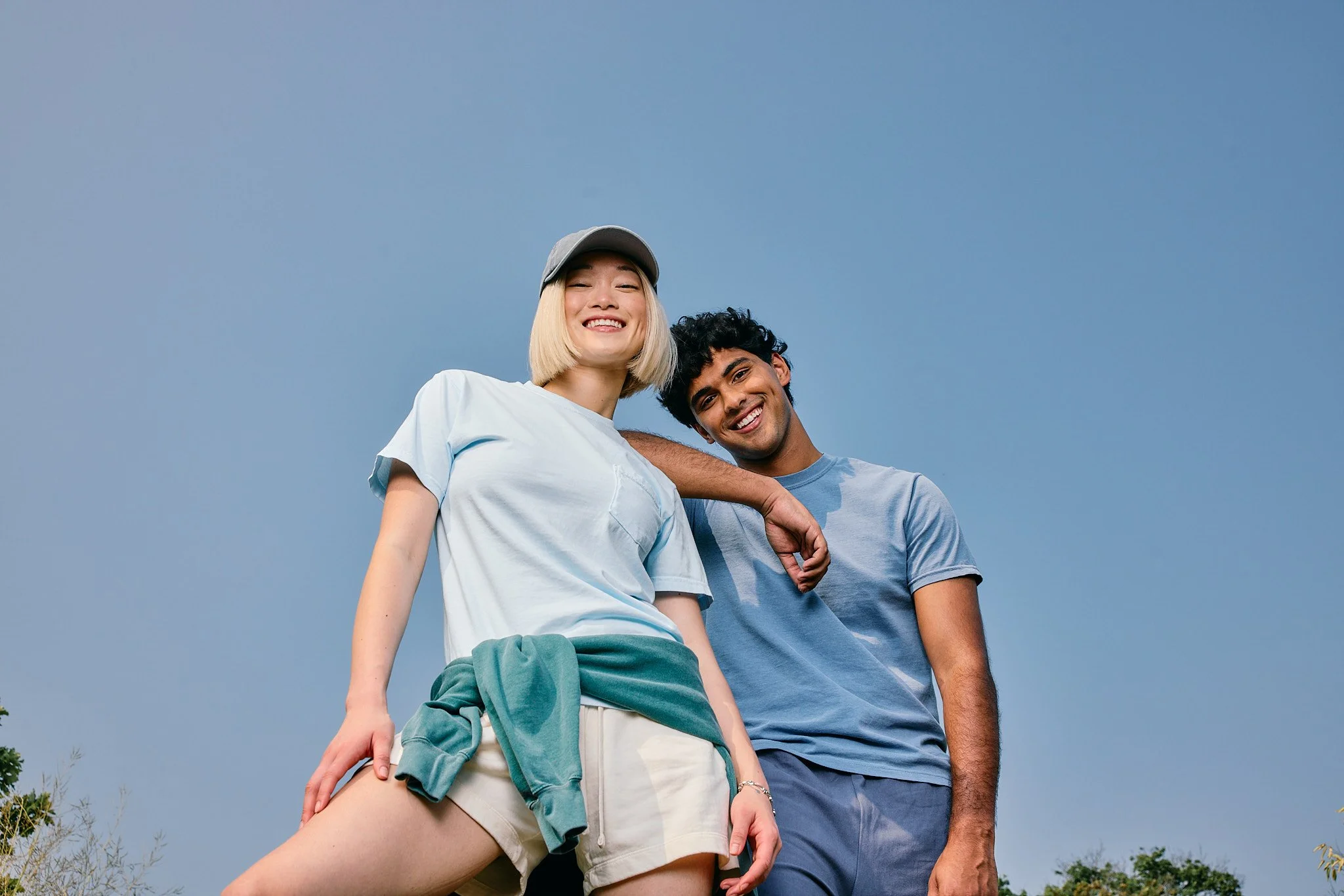 Smiling young woman with blonde hair wearing a gray baseball cap, light blue t-shirt, and shorts, standing outdoors with a young man with dark hair and brown skin, both under a blue sky.