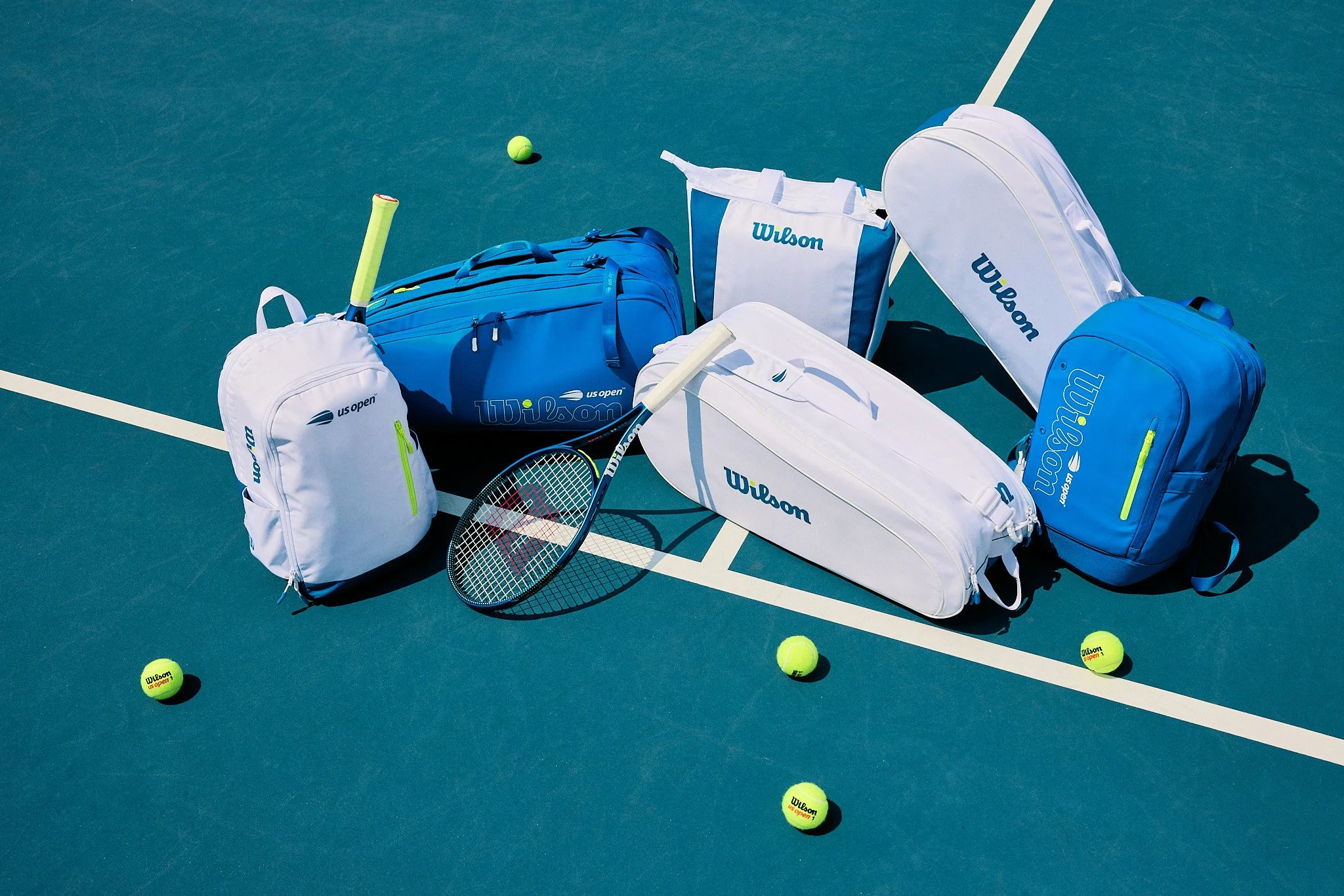 Wilson tennis bags and rackets on a tennis court with tennis balls scattered around.